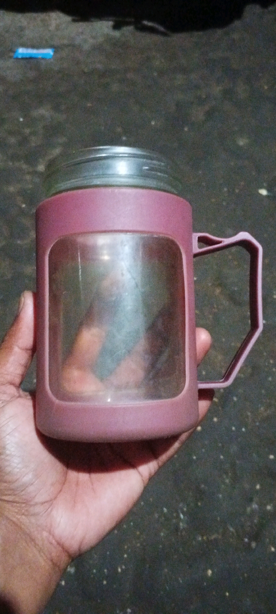 A person is holding a pink mason jar mug with a clear window on a dark, gravelly surface. The mug has a silver lid and a pink handle. The background is out of focus, but appears to be an outdoor setting. There is a blue object on the ground to the left of the mug.