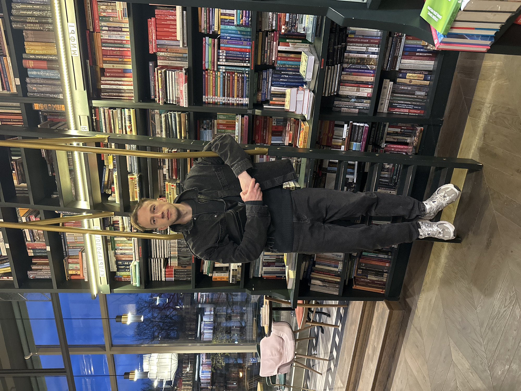 The image captures a man standing on a ladder in what appears to be a library or bookstore, potentially located in Saint Petersburg, Russia. He is dressed in black jeans, a black denim jacket, and white and silver sneakers. The man is positioned in front of bookshelves filled with a variety of books. He seems to be posing for the photograph.
The library or bookstore has a high ceiling and large windows that provide a view of the exterior, showing the blue sky and the dark shapes of trees. Seating is visible near the windows.
The books on the shelves cover a wide range of subjects, including history, culture, and literature. The shelves are organized and well-stocked. The presence of Russian text on many of the book covers indicates the likely location.