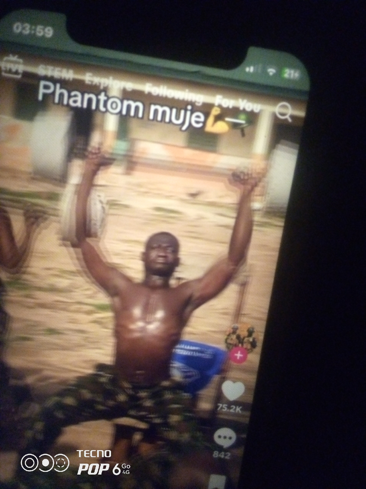 The image displays a smartphone screen, likely a video-sharing platform interface. The time "03:59" is visible in the top left corner, suggesting the video is playing at this time. The interface shows "STEM Explore Following For You" text. A username "Phantom muje" with a bicep emoji is prominent. Below, a shirtless Black man with dark skin is captured mid-action, with his arms raised above his head, his torso glistening. He is wearing camouflage pants. Behind him, a blue bucket and two other figures in military-like uniforms are partially visible. The background appears to be an outdoor, sandy or dirt area with some vegetation. On the right side of the screen, interaction icons are present: a red plus sign, a white heart with "75.2K" likes, and a speech bubble icon with "842" comments. At the bottom of the screen, the branding "TECNO POP 6 4G" is visible. The image is blurry due to the low resolution and possibly motion blur. There is no information in the image to determine its specific location in Howayi, Nigeria.