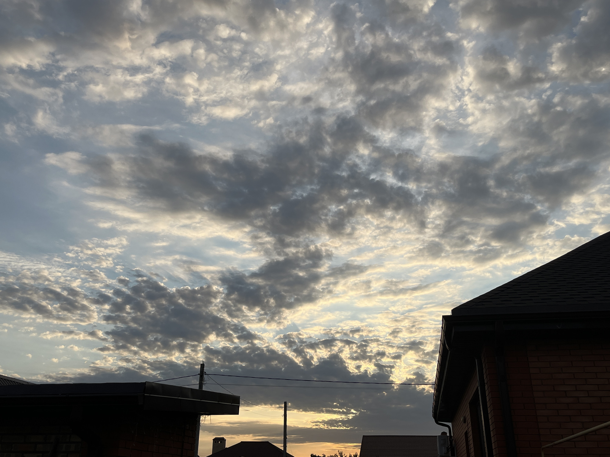 A low-angle shot captures a dramatic sky filled with a mix of dark and light clouds, some illuminated by a warm sunset. Silhouetted against the sky are the rooftops of brick buildings in Taganrog City District, Russia. The foreground features the dark, angular shapes of building eaves and tiled roofs. Electrical wires and a utility pole are visible, adding to the urban landscape. The lighting suggests it is late evening or early morning.