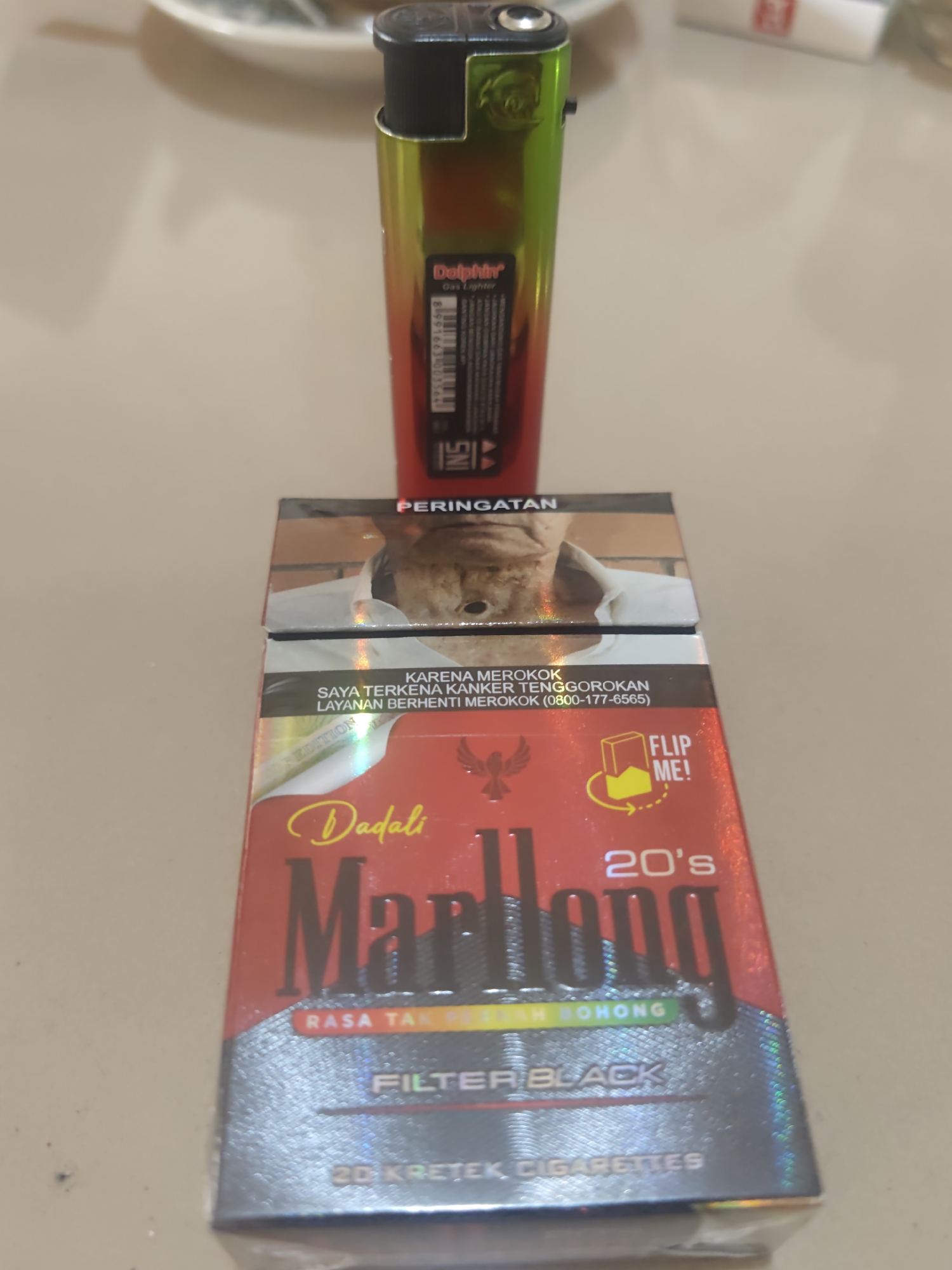 A pack of Marlboro 20's Filter Black kretek cigarettes, with an image of a person on the front and a warning in Indonesian, sits on a light-colored, textured surface, possibly a table. The pack is partially covered by a multi-colored lighter, which has a brand name "Dolphin" and "Gas Lighter" printed on it. The background is blurred and shows a white plate and a portion of a cigarette pack with red lettering. The scene appears to be indoors in Pematang Reba, Indonesia.