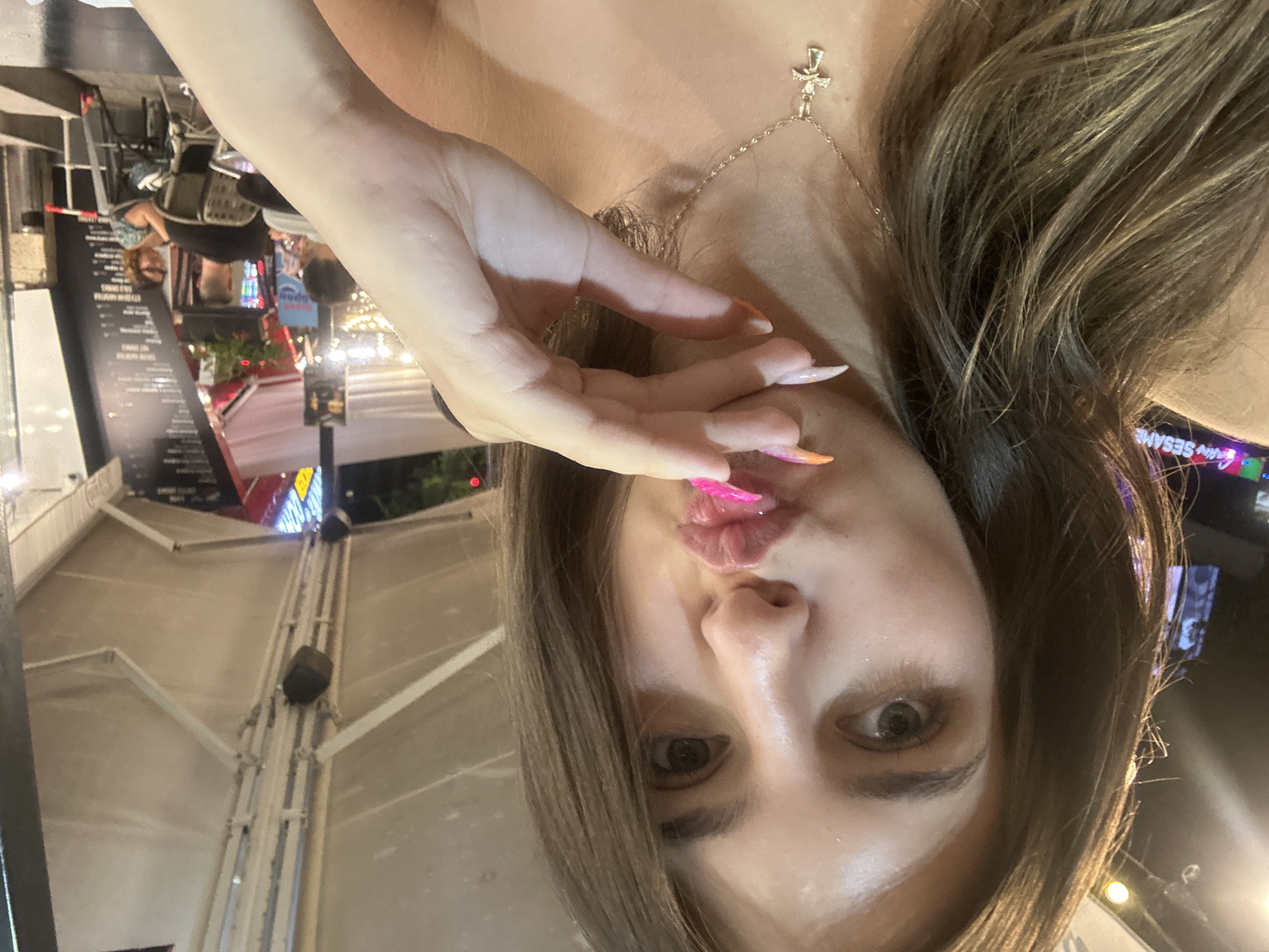 A woman with long, brown hair and striking makeup is taking a selfie. She is leaning into the camera, puckering her lips which are adorned with bright pink lipstick. Her right hand is raised, touching her chin and drawing attention to her meticulously manicured nails. She is wearing a delicate gold necklace with a cross pendant. The background suggests an outdoor seating area of a restaurant or cafe at night. Visible in the distance are other patrons seated at tables, illuminated by ambient lighting and neon signs. One sign reads "GRAN SESAME." The overall ambiance is casual and lively, typical of a bustling city evening. The location is identified as the city of Varna, Bulgaria.