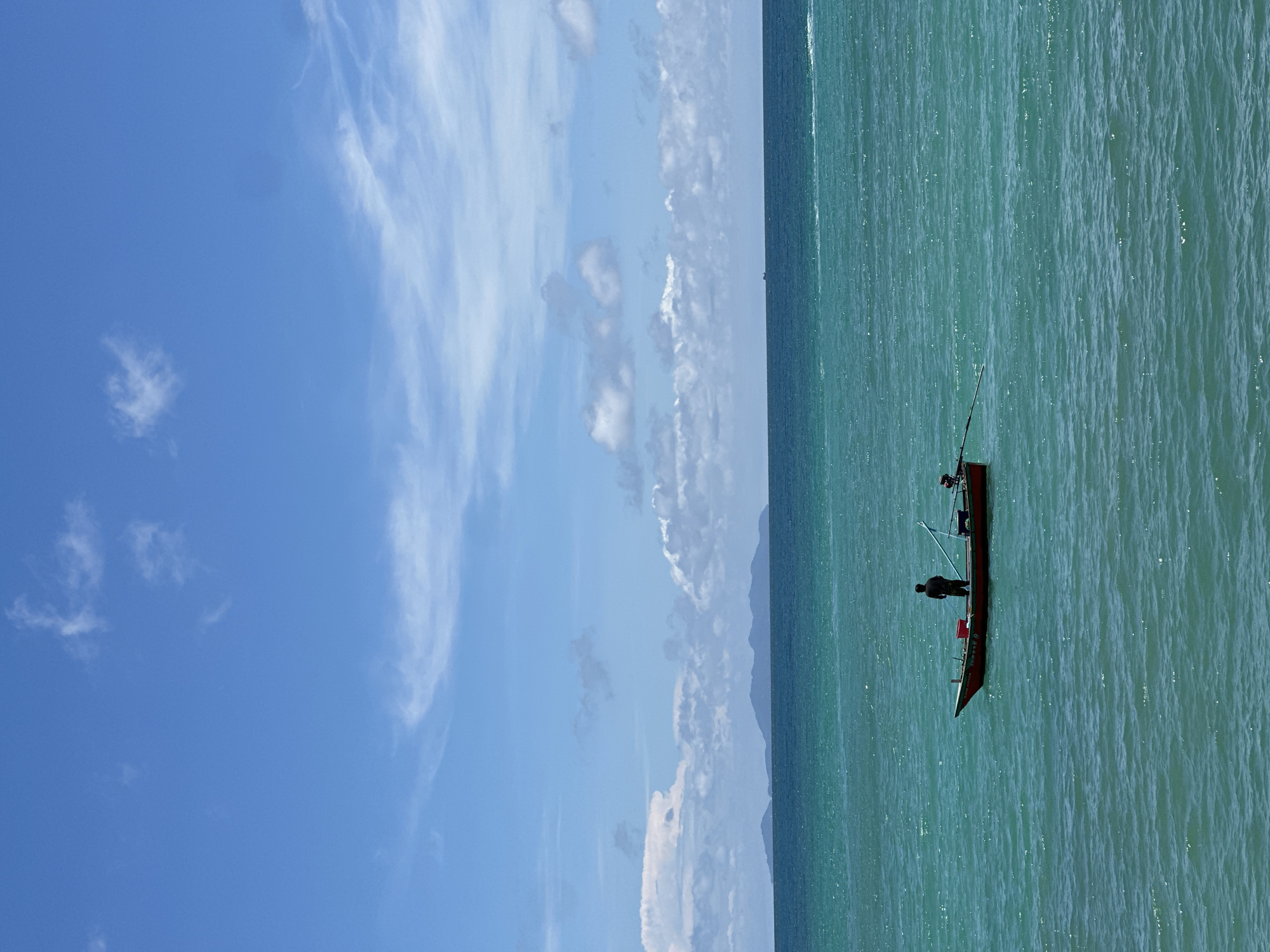 A lone figure stands in a small red boat on a calm, turquoise sea. The boat, with an outboard motor attached, is positioned in the middle ground of the frame. In the background, a faint, hazy silhouette of islands can be seen against the horizon, under a bright blue sky dotted with scattered white clouds. The scene suggests a serene, possibly remote, coastal location.