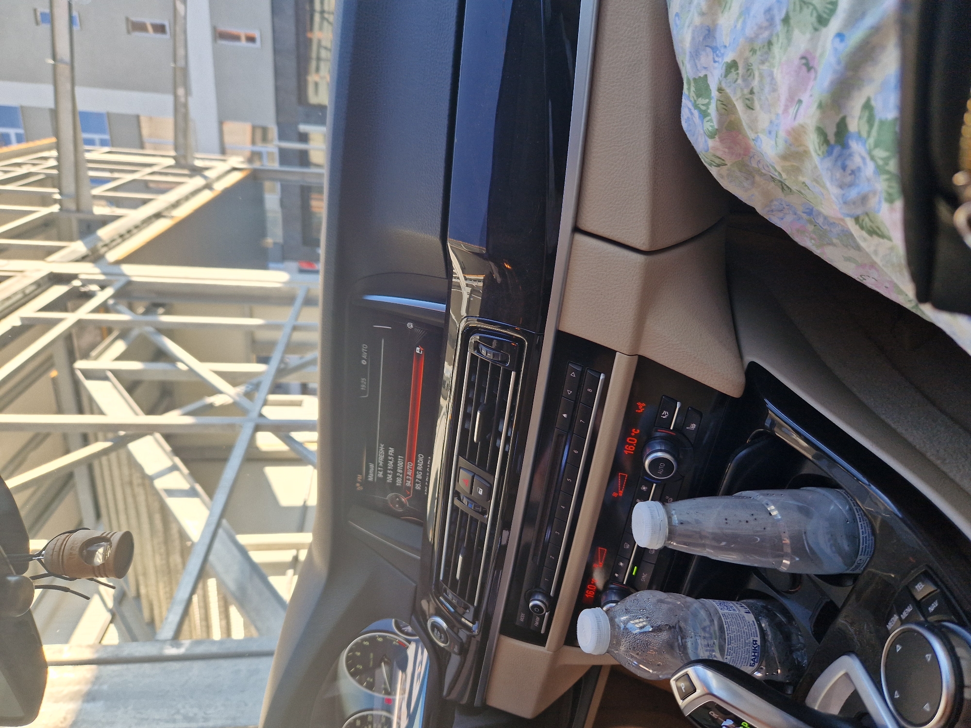 An interior shot of a car cabin, taken from the passenger seat. The dashboard features a digital display showing radio stations and climate control settings with a temperature of 16.0°C. Two plastic water bottles are visible in the center console, one partially obscured. A floral-patterned fabric is draped over the passenger's lap. Outside the car, a modern building and scaffolding are visible through the windshield, suggesting an urban environment. A small, hanging air freshener dangles from the rearview mirror.