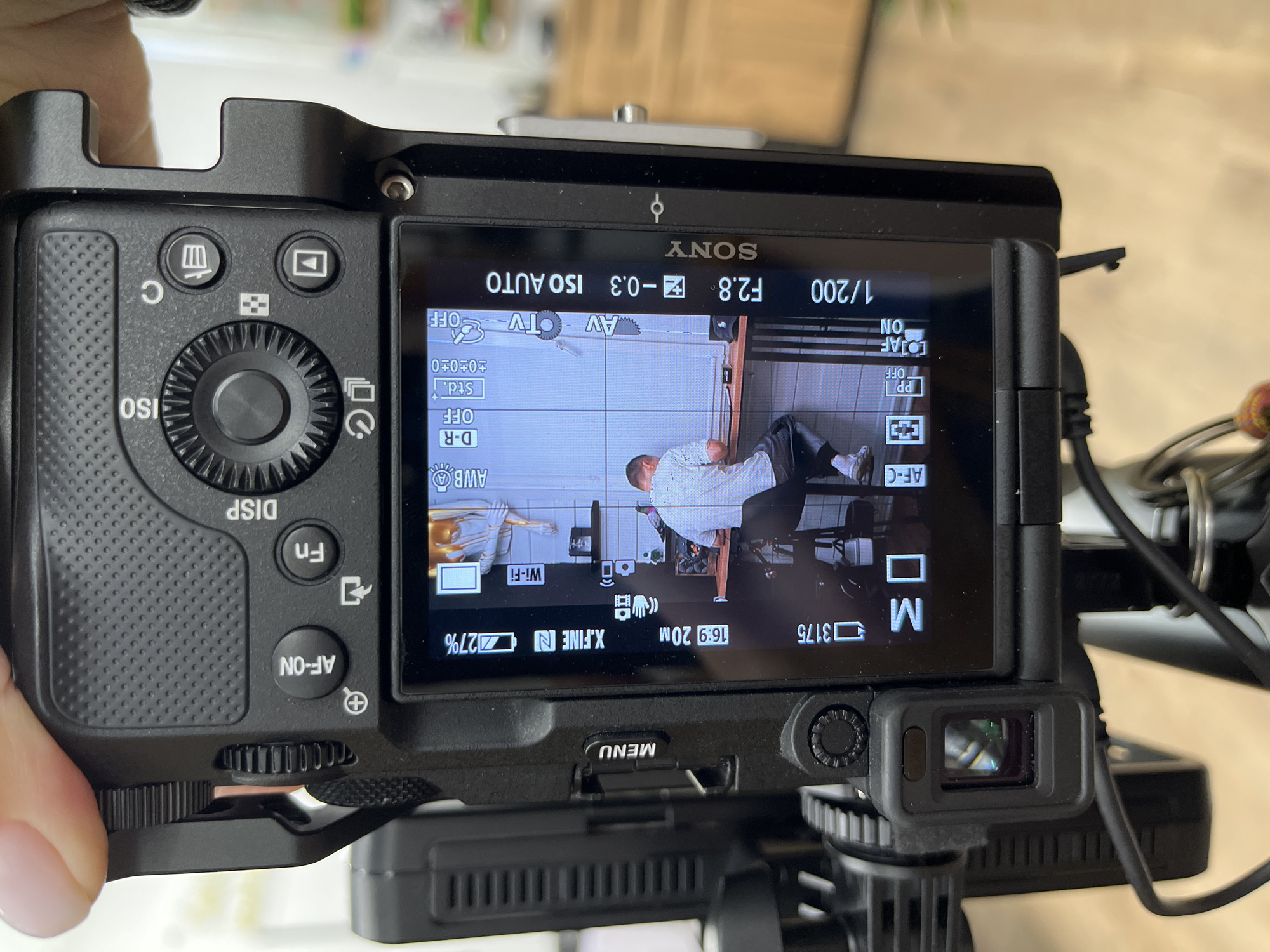 The image shows the back of a Sony digital camera with its LCD screen active, displaying a live view of a person sitting at a desk, likely filming or photographing themselves. The camera is held by a person's hand from above. The screen displays various camera settings, including aperture (F2.8), shutter speed (1/200), ISO (ISO AUTO), and a battery level indicator at 27%. The environment visible on the screen appears to be an indoor setting with a white tiled wall behind the person and a wooden desk. The camera itself has several buttons and dials visible, including "AF-ON", "Fn", "DISP", and a directional pad. The exterior of the camera shows a textured grip and a viewfinder. The image was taken in Chaikovsky, Russia.