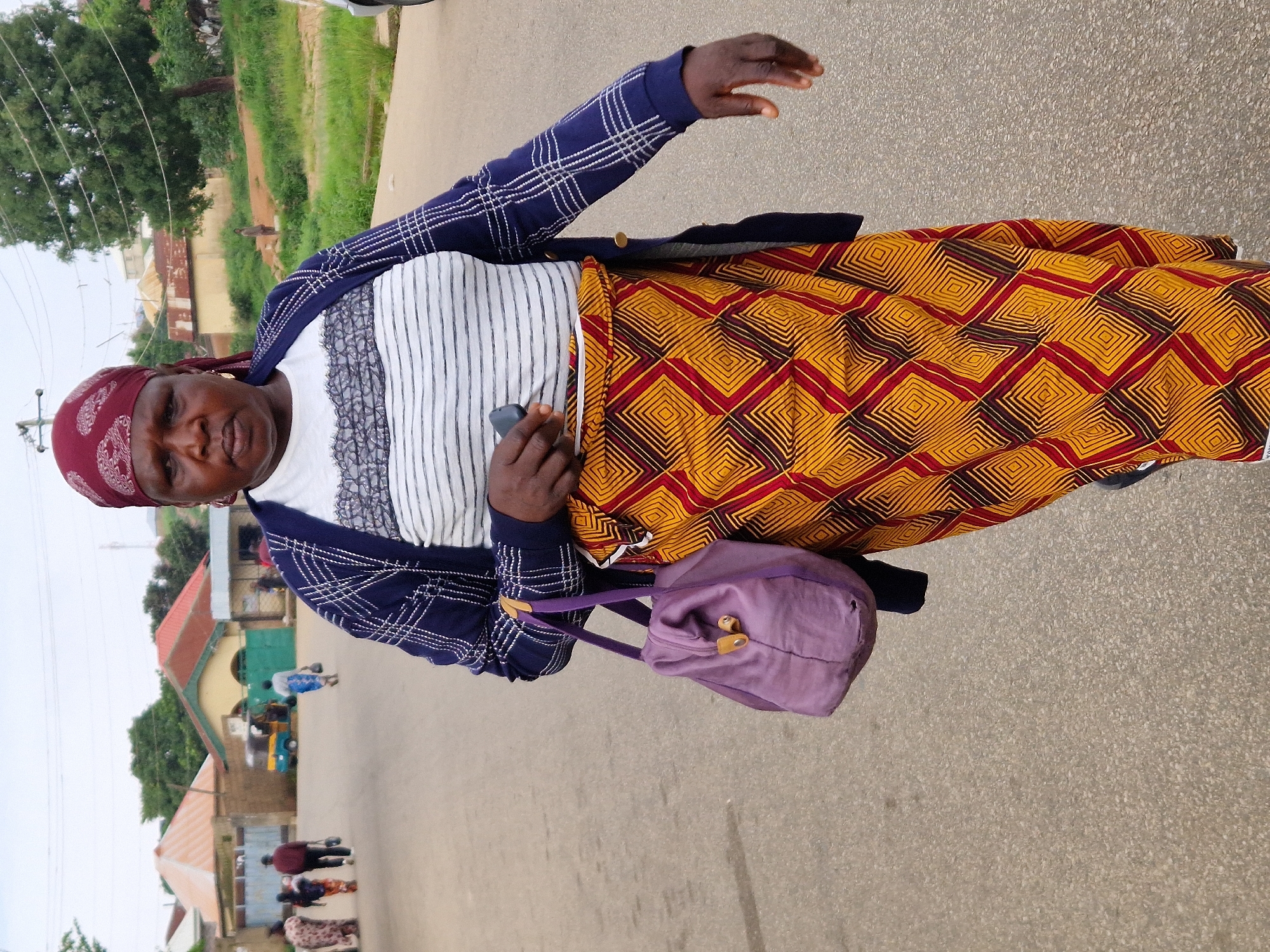 In Kabong, Nigeria, a woman walks along an asphalt road under an overcast sky. She wears a burgundy headwrap, a white and black striped shirt, and a blue cardigan with white plaid detailing. Her lower body is adorned with a vibrant, patterned wrap skirt featuring diamond shapes in shades of yellow, red, and brown. A purple handbag hangs from her left shoulder. She holds a dark-colored object, possibly a phone, in her right hand. In the background, several buildings with red and orange roofs are visible, along with a blue and yellow auto-rickshaw and other pedestrians walking away from the camera. Green vegetation lines the side of the road, suggesting a semi-urban or suburban setting.