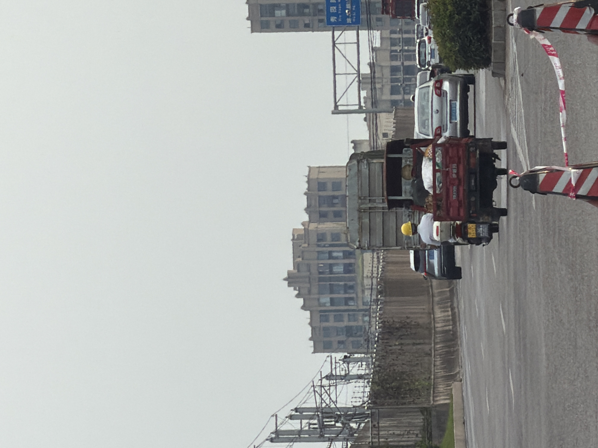 In Jiangjin, China, on a cloudy day, a busy city street scene unfolds. A three-wheeled vehicle, loaded with goods and with two individuals aboard, is central to the image, moving in the foreground. Behind it, a silver sedan navigates the traffic. To the right, a motorcycle with a rider wearing a yellow helmet is also visible. Further in the background, several multi-story buildings stand, one of them appearing to be a large, older structure with a distinct architectural style. A traffic light pole with a blue sign hangs over the road. The scene is characterized by moderate traffic and a general sense of urban activity.