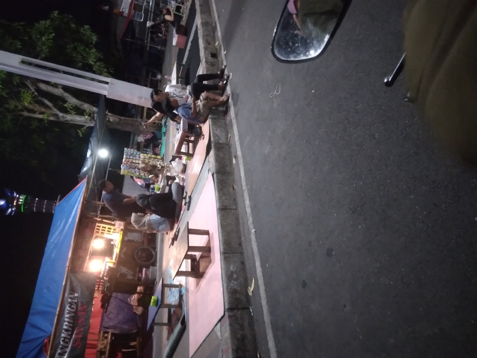 The image depicts a nighttime scene at an outdoor food stall in Purwokerto, Indonesia, likely a street food vendor selling Indonesian specialties. A blue tarp covers the stall, illuminated by warm lights, and a banner with the words "NGKRINGAN SAN-SAN" is visible. Several people are seated on the ground or on small stools around low tables. A man in a dark shirt is standing and appears to be attending to the stall. Two children and adults are gathered around a table. In the foreground, a motorcycle mirror reflects a partial view of the scene and the rider. The setting appears to be alongside a paved road with a curb, suggesting a roadside establishment.