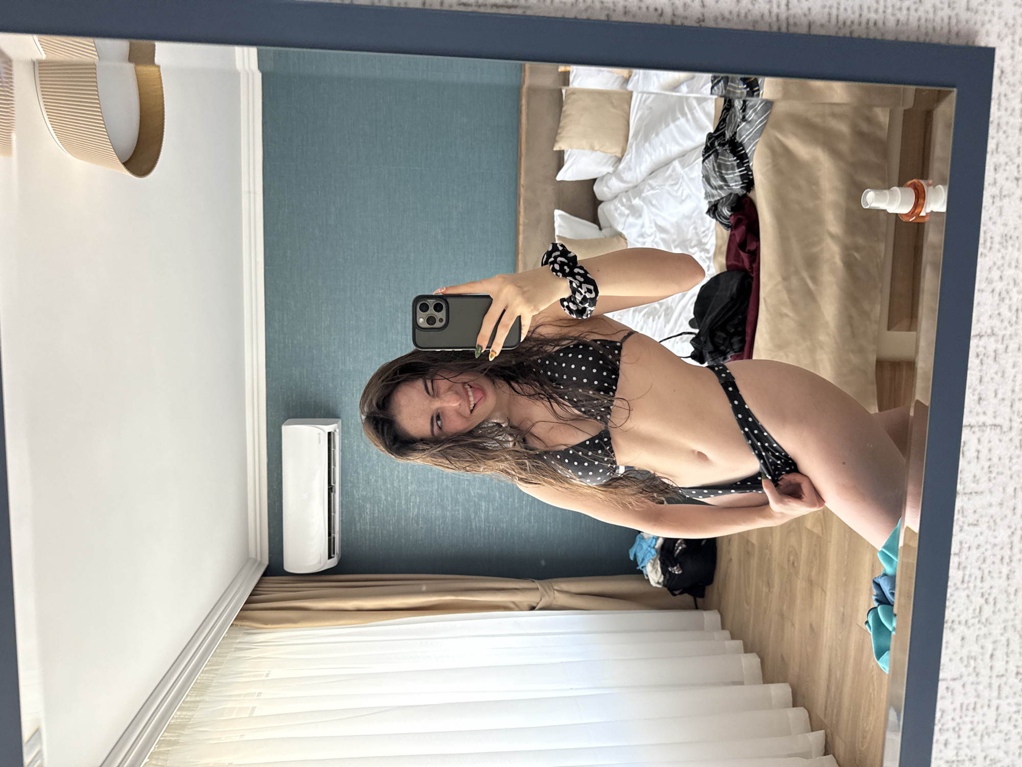 A young woman with wet, dark hair is taking a mirror selfie in a bedroom. She is wearing a black and white polka dot bikini and winks at the camera while sticking out her tongue. Her right hand holds a smartphone, and a black and white scrunchie is on her left wrist. The room has light wood floors, a turquoise accent wall, and a large bed with white linens in the background. A white air conditioning unit is mounted on the wall. Curtains are visible on the left side of the frame. A bottle of white spray is on the floor in the foreground. The overall impression is of a casual, personal moment in a hotel room, possibly in Sveti Vlas, Bulgaria, given the context.