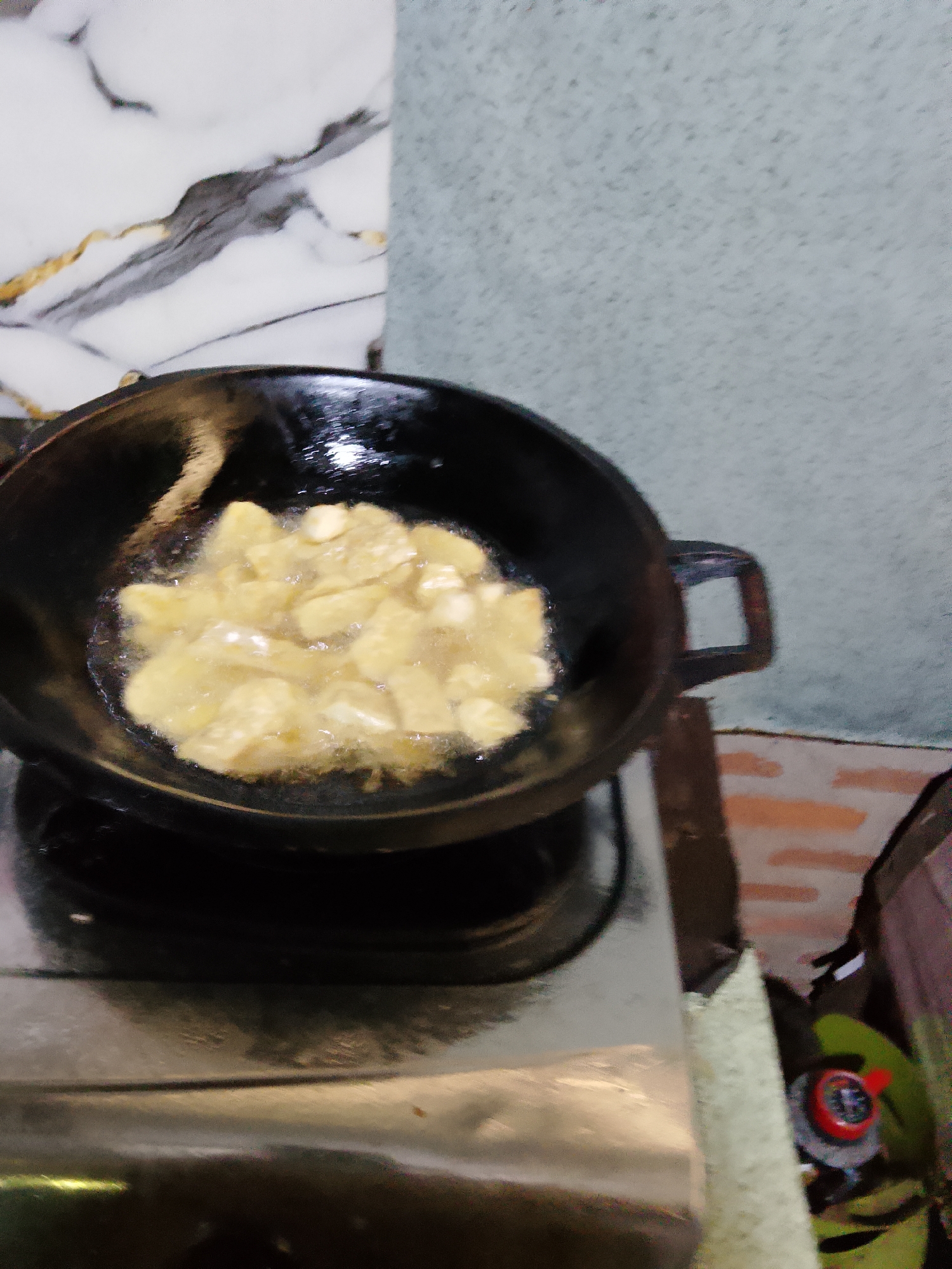 A black wok filled with golden-brown potato slices being fried in oil sits on a gas stove. The wok has a handle on the right side. The background is a wall with a marble-like pattern on the left and a textured, pale grey surface on the right. A gas burner is visible beneath the wok. The scene appears to be in a kitchen.