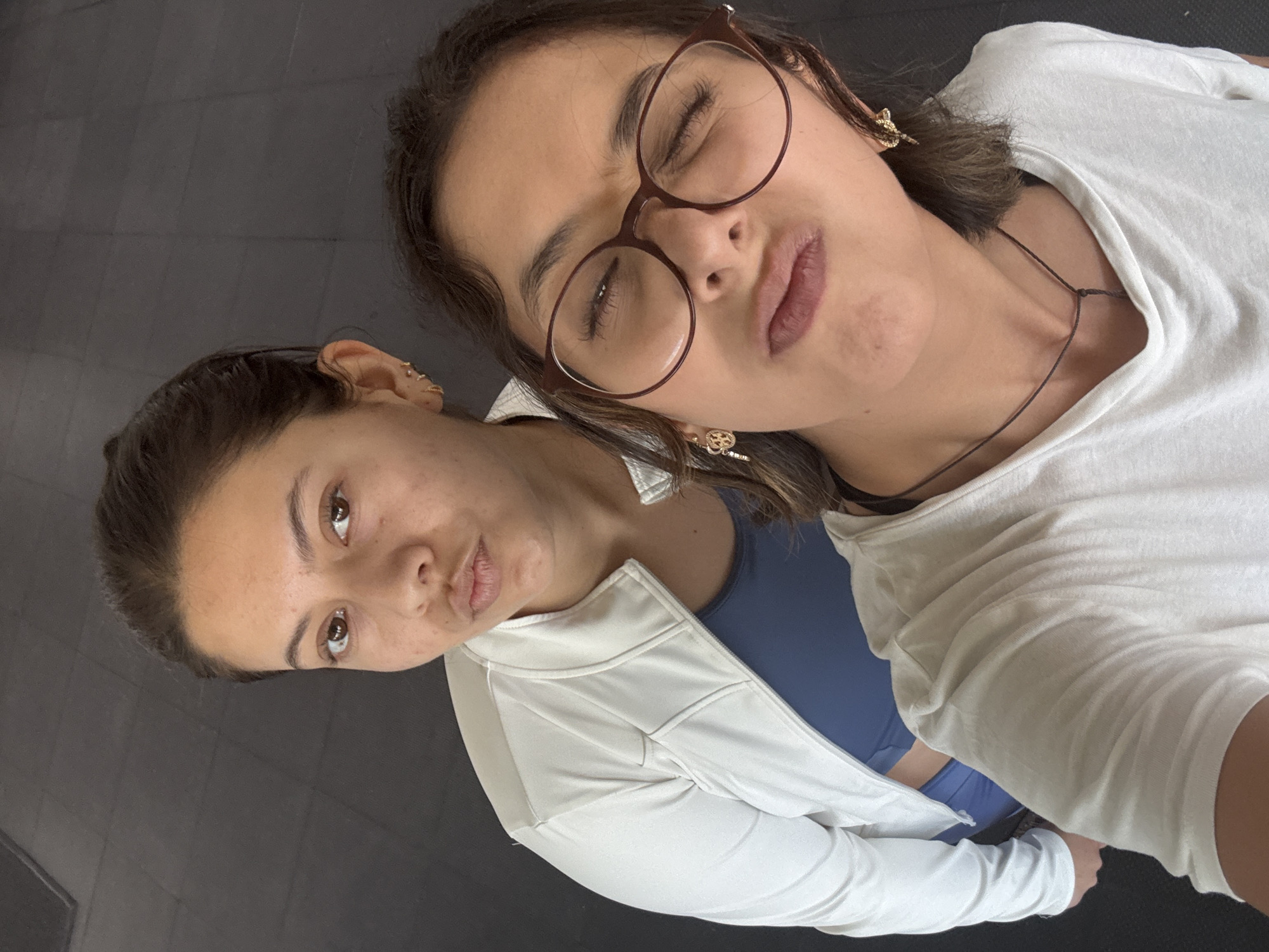 Two young women pose for a selfie in San Andrés Cholula, Mexico. The woman on the right wears round glasses, a white t-shirt, a necklace, and small gold earrings. Her eyes are partially closed, and she is making a kissing face. The woman on the left has her hair pulled back and is wearing a blue sports bra under a white zip-up jacket. She is also making a kissing face and looking directly at the camera. The background is a plain, dark surface, likely a floor.