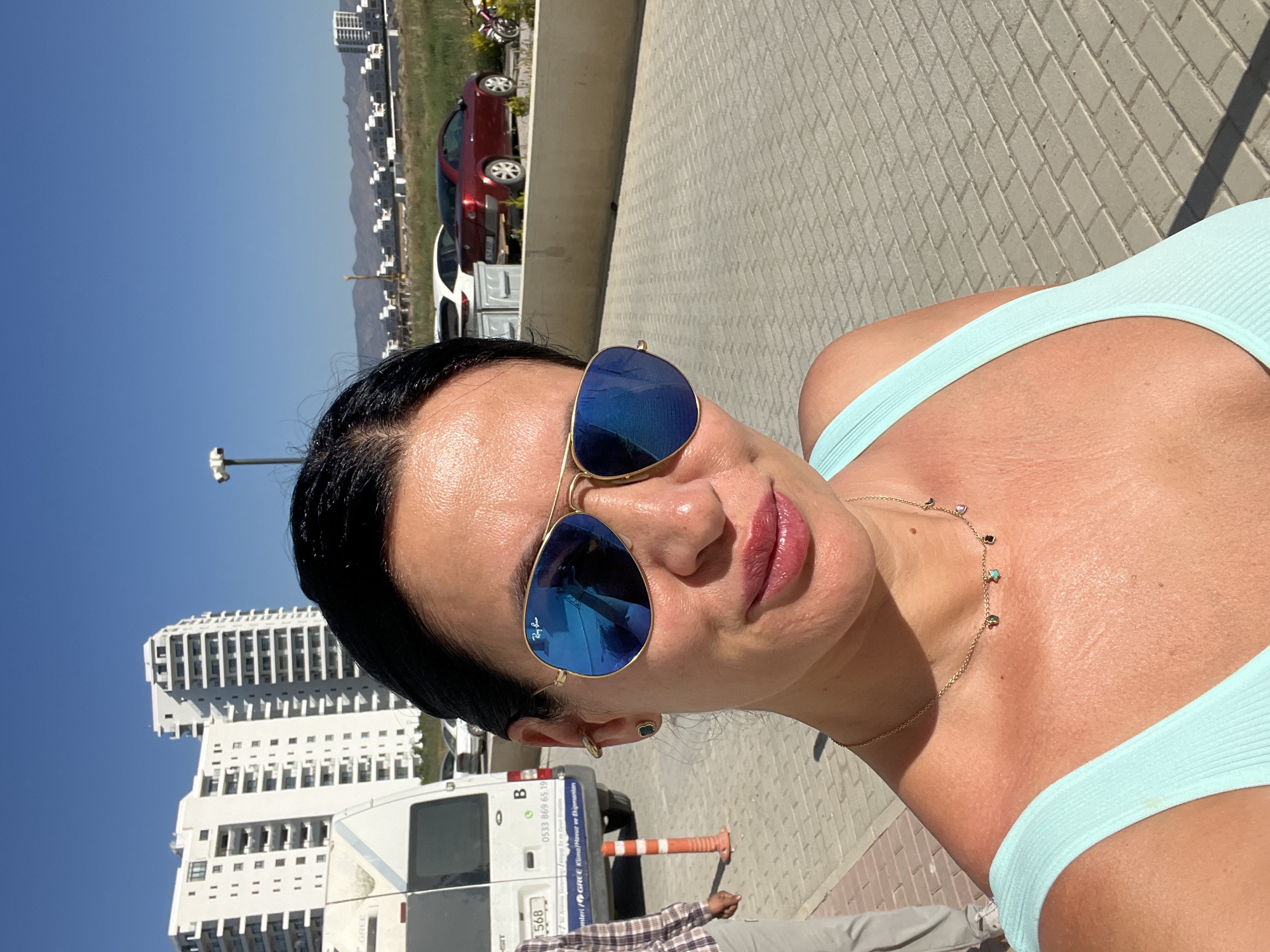 A woman is captured in a sunlit outdoor portrait, tilted to her right. She has dark hair and wears large, reflective blue aviator sunglasses, displaying the "Ray-Ban" logo. Her lips are glossy, and she wears a light blue ribbed top along with a delicate gold necklace adorned with small, colorful charms like stars and moons. A small black clover-shaped earring is visible.
The scene appears to be a paved area, likely a sidewalk or street. In the middle ground, a white Ford Transit van is prominent, featuring Turkish text including "GREE Klima Havuz ve Ekipmanları" (GREE Air Conditioning Pool and Equipment), "0533 869 65 19", "Aritme Sisteme", "1568", and the letter "B". Partially visible behind the van is a person's arm and lower torso, wearing a plaid shirt and grey pants. A traffic cone is also seen near the van.
The background reveals several modern, multi-story white buildings under a clear blue sky. A red car and other vehicles are parked further down the paved area, with mountains visible in the distant horizon. The bright light suggests it is daytime, with strong shadows indicating intense sunlight, consistent with the provided location context of Trikomo, Northern Cyprus.
