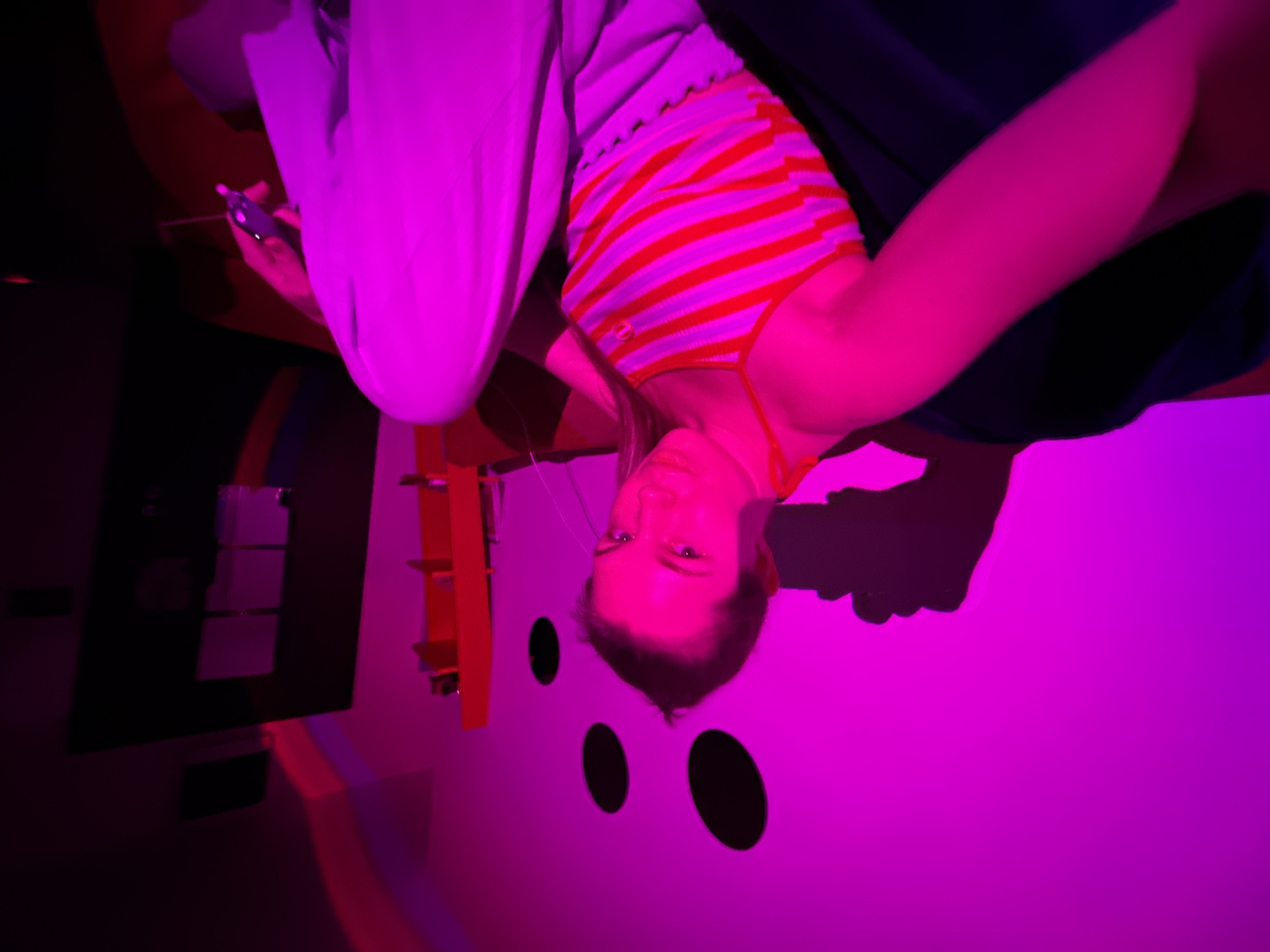 The scene depicts an indoor room illuminated by a prominent magenta light source, located within what is indicated as the city of Vozdovac Urban Municipality, Serbia. A single individual, appearing female, is seated centrally, facing the camera directly with a neutral expression. She wears a light-colored top with horizontal orange stripes and light-colored wide-leg pants. Her right arm is extended forward, holding a dark-colored object, likely a smartphone, which casts a distinct shadow on the wall to her left. A thin, dark cable is visible near her left arm. The background wall, rendered magenta by the lighting, features three dark, circular decorative elements. To the right, an orange shelving unit is visible, adjacent to a dark, vertical rectangular panel or door that contains lighter, reflective rectangular sections. A faint, curved red light streak is discernible on the upper right wall surface. The individual is positioned on a dark-colored seating surface. Specific geographical coordinates or building details within Vozdovac are not discernible.