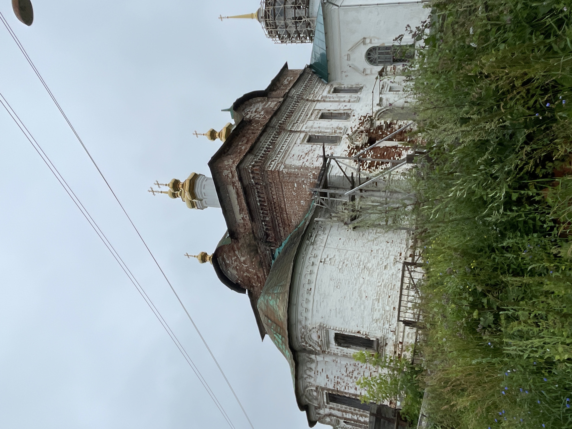 An outdoor, low-angle, medium shot shows an old, white brick church with a red brick upper section, possibly under construction or renovation, in the city of Kstynino, Russia. The church has several golden domes topped with crosses. Scaffolding is visible on the side of the building. The church is surrounded by overgrown green grass and foliage, with a grey, overcast sky above. Power lines are visible in the upper portion of the image.