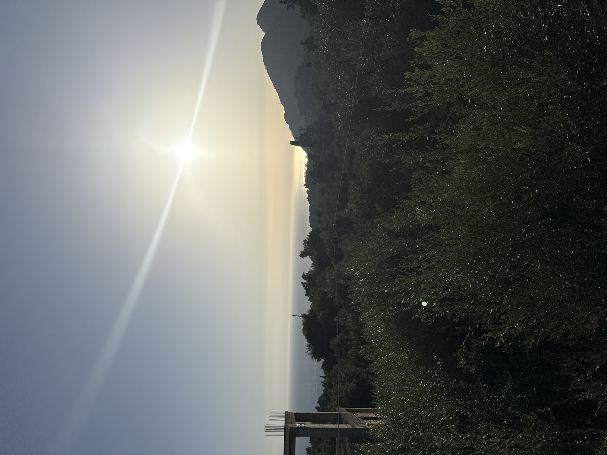 An outdoor, eye-level shot captures a scenic view of the sea and a mountainous landscape under a bright, hazy sun. The sun is positioned in the upper right quadrant of the image, emitting a strong glare that extends across the sky. A bright, linear light streak emanates from the sun, diagonally crossing the frame. The sea is visible in the midground, appearing calm and stretching towards the horizon. To the right of the sea, a silhouette of a mountain range is prominent, with its peaks softened by the haze. In the foreground, dense, dark green foliage, likely olive trees, dominates the lower half of the image. To the left of the frame, a partially constructed concrete building stands unfinished, its rebar protruding from the top. The scene is set in Kokkini, Greece.