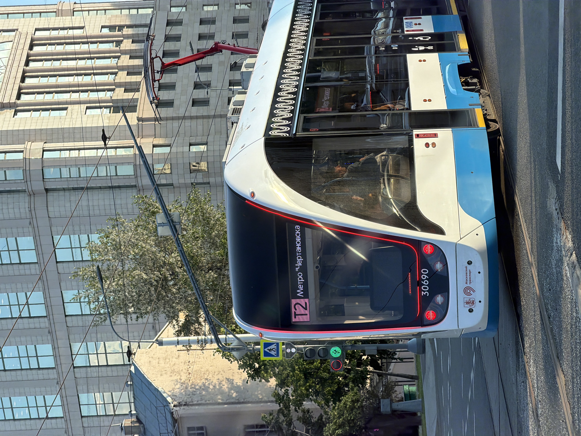 This image depicts a modern, white and blue tram in an urban setting in Moscow, Russia, captured during bright daylight. The tram, a central subject, is stationary on its tracks, likely paused at a traffic light, which displays a red signal for trams.
The tram's front explicitly shows its route as "T2" and destination "Метро "Чертановская"" (Metro "Chertanovskaya"). Its registration number, "30690," is visible on the lower front. The "Московский Транспорт" (Moscow Transport) logo and text are also present on the front lower panel. Along the upper side of the tram, a distinctive wavy, decorative pattern is illuminated. Through the large windows, several passengers can be seen inside, some seated and others standing, and a reflection of a person is visible in the front window. Another internal display within the tram indicates "Metro Novogireevo" (Метро "Новогиреево").
In the background, a tall, modern building with a grid of numerous windows rises prominently. Overhead power lines, essential for the tram, crisscross the scene, and a small, dark bird is perched on one of these wires. Lush green trees, some appearing to be in blossom, provide a touch of nature against the city backdrop. A pedestrian crossing sign and traffic lights are also visible, indicating an active street environment. The scene is well-lit by sunlight, suggesting a clear morning or afternoon.