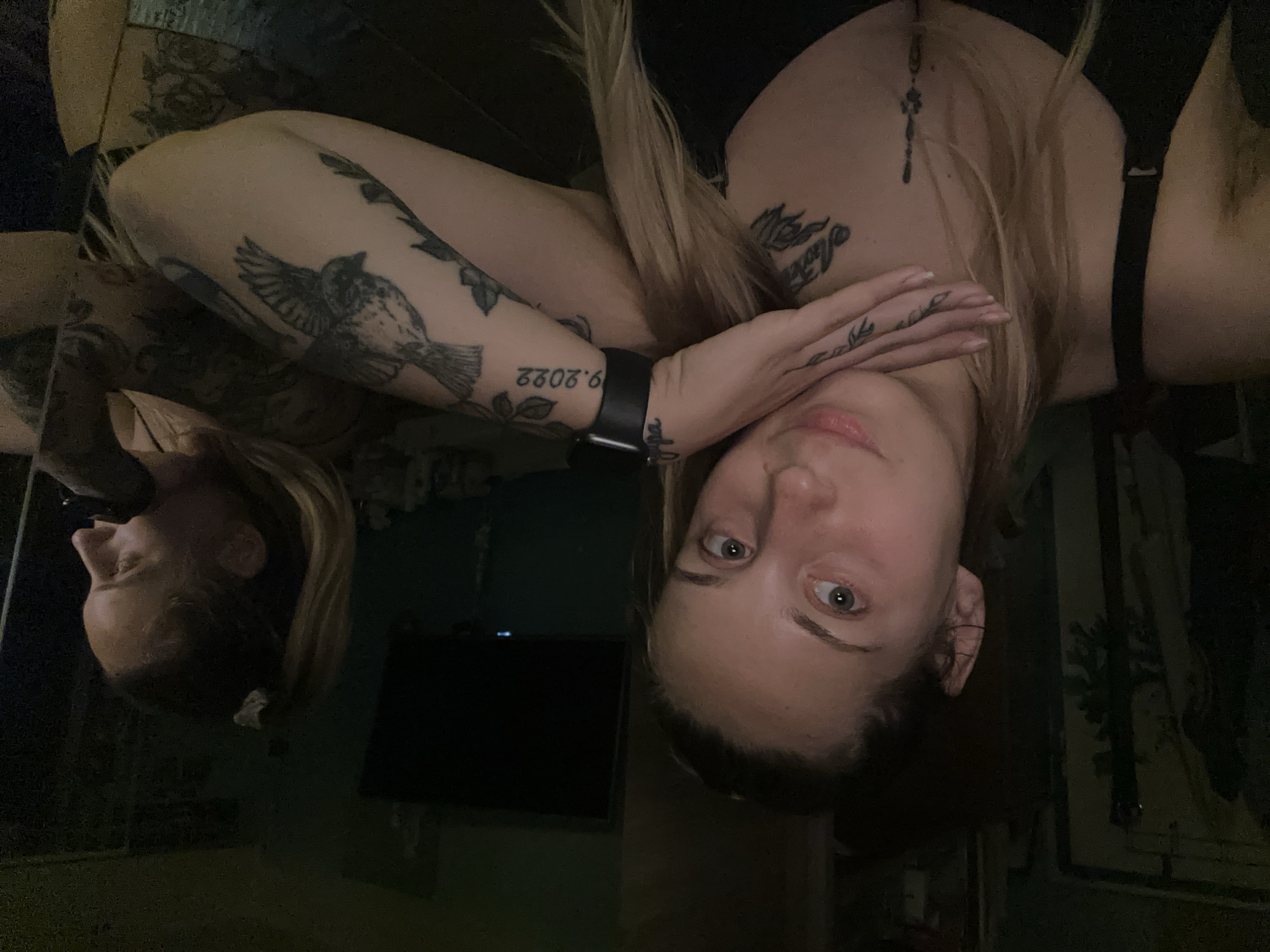 Two women are in a dimly lit room. The woman in the foreground is looking directly at the camera, with her left hand resting on her chin. She has blonde hair and is wearing a black bra. She has several tattoos on her arm and chest. The woman in the background is in profile, looking to the right. She also has blonde hair, pulled back into a ponytail, and is wearing a black top. The room appears to be a living space, with a television visible on the wall in the background. The overall impression is that of a casual, private setting.