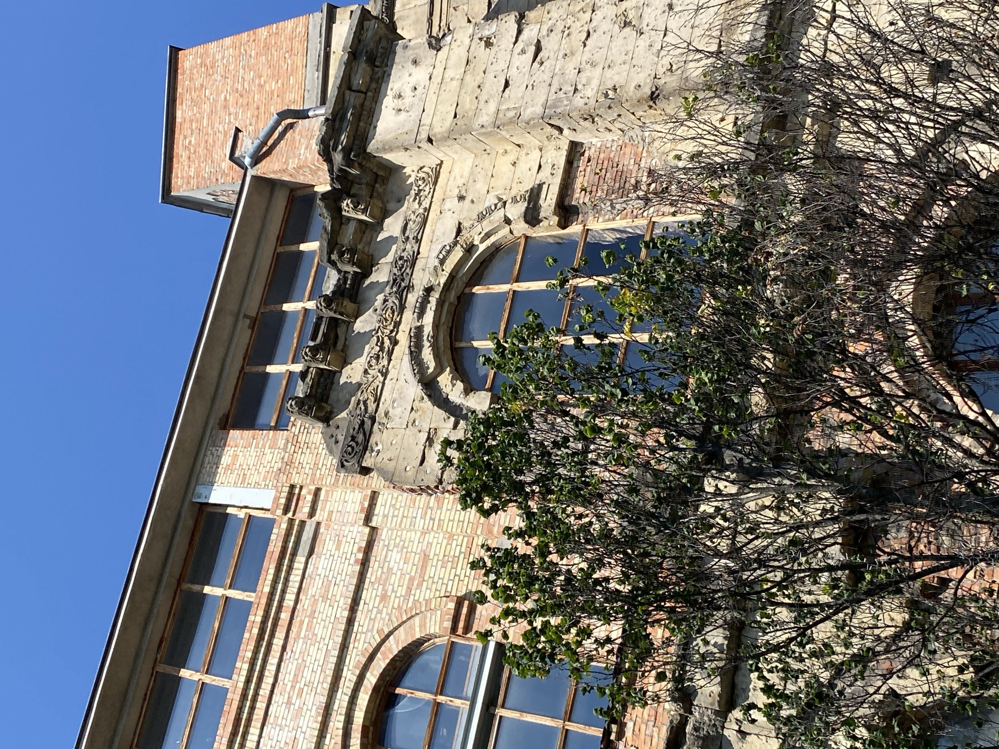The image shows the exterior of a weathered brick building, likely in Warsaw, Poland, under a clear blue sky. The building exhibits signs of age and possibly past damage, with visible pockmarks on the stone facade and some exposed brickwork. Architectural details include arched windows with wooden frames and glass panes reflecting the sky, as well as decorative stone carvings along the upper edge of the building. A tall brick chimney extends from the roof on the right side, with a downspout attached. In the foreground, the branches of a tree partially obscure the building; some branches are bare, while others are covered in green leaves. The perspective is from below, looking up at the structure. There are no people or active events depicted in the scene.