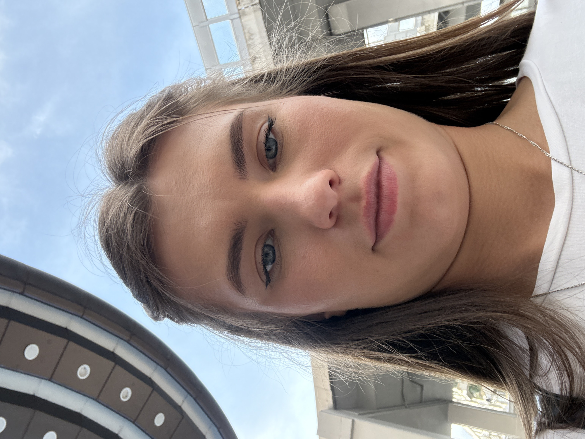 A close-up, low-angle selfie of a young woman with brown hair and blue eyes is captured outdoors. She is wearing a white t-shirt and a thin silver necklace. Her hair is partially pulled back, with strands framing her face. The background shows a bright blue sky with some clouds, and architectural elements including a curved structure with circular lights and a modern building with visible window frames. The location is Haizhu District, China.