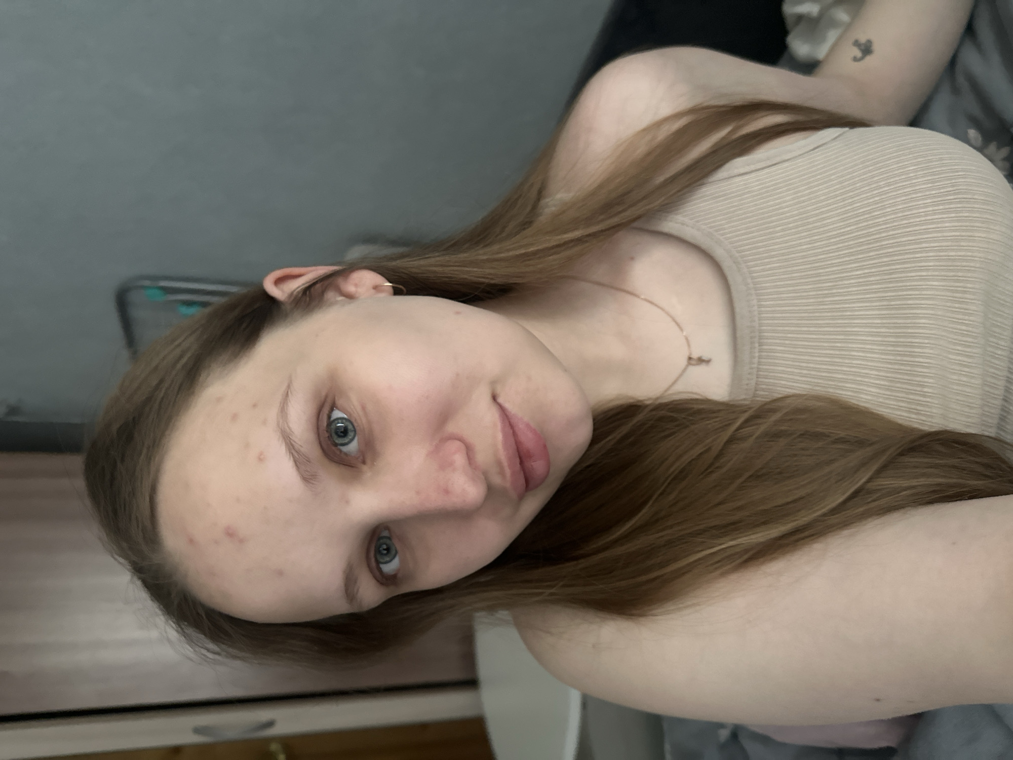 A young woman with light brown hair and blue eyes is the central subject of the image. She is fair-skinned and has some acne on her forehead and nose. She is wearing a light tan, ribbed tank top and a delicate gold necklace. A small tattoo is visible on her left arm. The background is blurry, but it appears to be a room with grey walls and a bed with grey and white bedding. The location in Chaikovsky, Russia cannot be determined from the image.