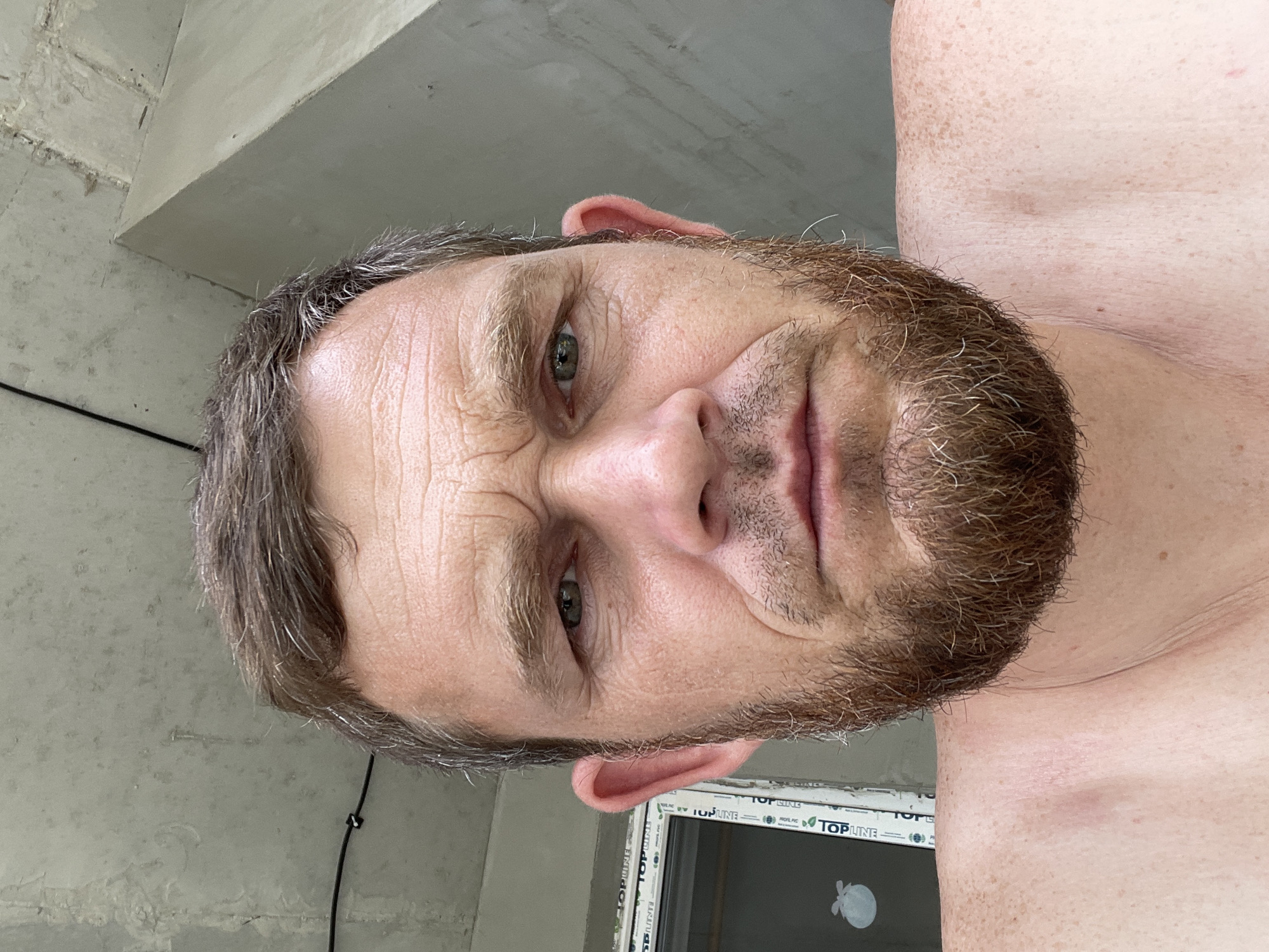 A man with a beard and short brown hair is looking directly at the camera. He has wrinkles on his forehead and around his eyes. His shoulders are bare, and he has freckles on his skin. Behind him, a window with the brand name "TOPLINE" is visible. The background also shows a concrete ceiling and wall, suggesting an indoor construction site. The image was taken in Krasnodar, Russia.