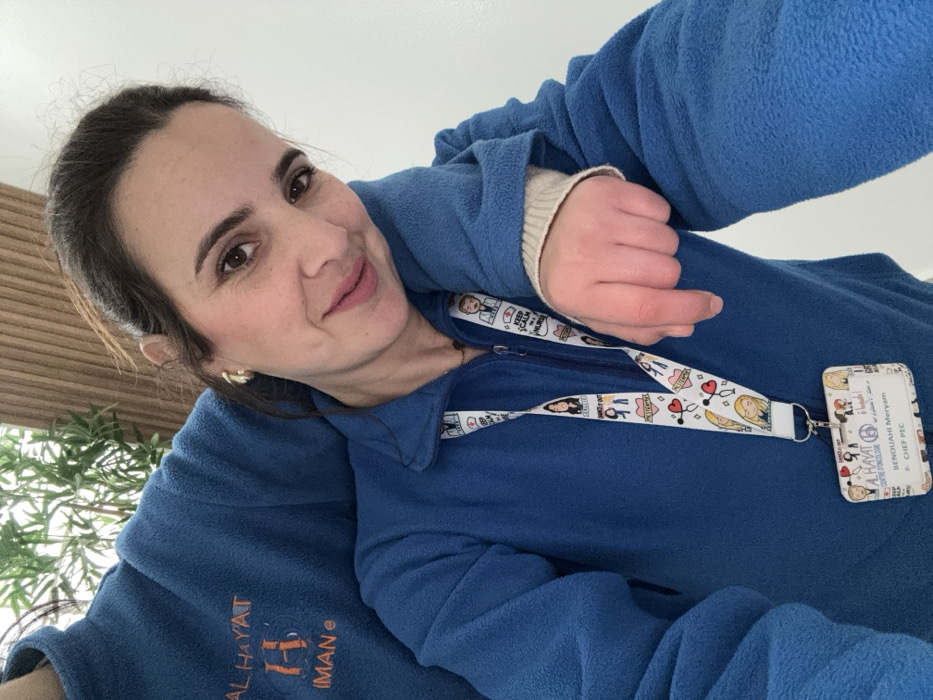 The image shows two individuals inside the Al Hayat Oncology Center in Meknes, Morocco. The foreground features a female with dark hair pulled back, wearing a blue fleece quarter-zip jacket, a gold-colored hoop earring in her right ear, and a faint smile directed at the camera. Around her neck is a white lanyard with colorful medical-themed illustrations, which holds an identification card. This ID card identifies her as "BENOUAHI Meryam" with the title "CHEF PEC" for "AL HAYAT CENTRE D'ONCOLOGIE". Partially visible behind her, primarily her left shoulder and arm, is a second individual also wearing a blue fleece garment. This garment has orange embroidery on the upper left shoulder area, reading "AL HAYAT", a stylized "H", and "IMAN". The second individual's left arm is draped over the primary subject's left shoulder. The background consists of a light-colored wall on the left, a wooden slat wall on the right, and green foliage, indicative of an indoor setting.
