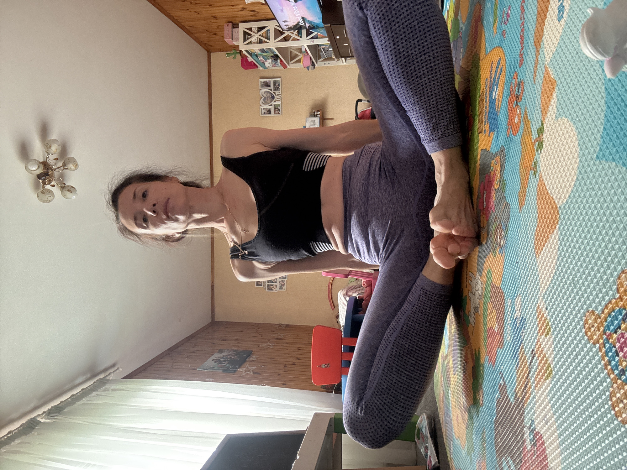 A woman is sitting on a colorful play mat in a room. She is wearing a black sports bra and purple leggings. Her legs are bent and her feet are touching. She is looking up at the camera. The room has wood-paneled walls and a white ceiling with a light fixture. There are some children's toys and furniture in the background, including a red chair and a shelf unit. A window with sheer curtains is visible on the left side of the image.