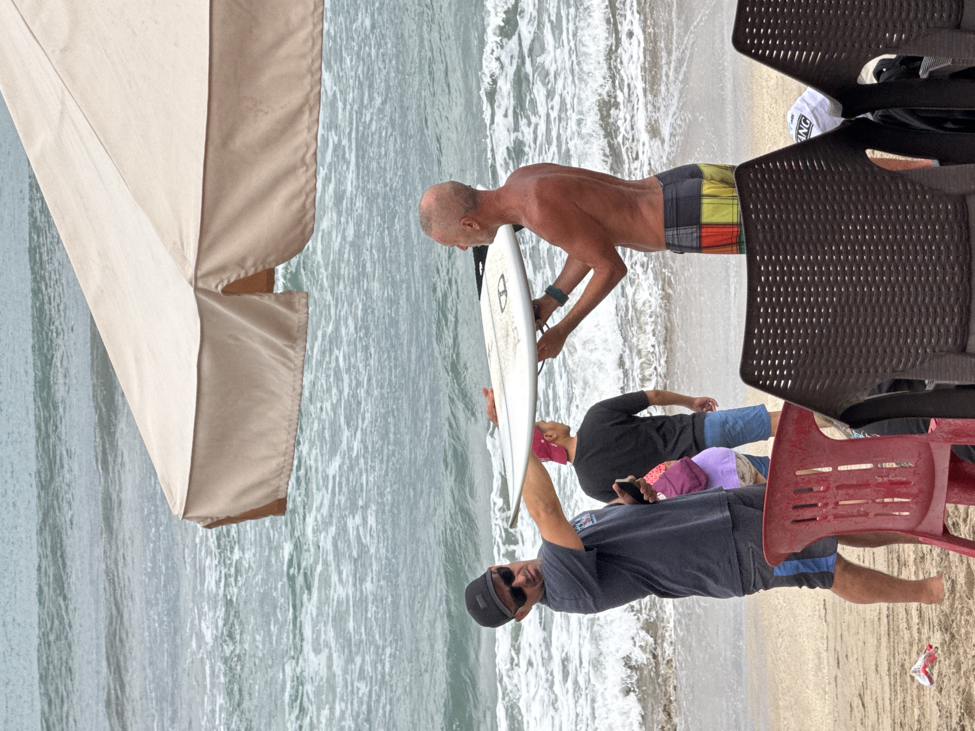 Two men are on a sandy beach in Kuta, Indonesia, by the ocean. The man on the left is wearing sunglasses, a dark t-shirt, and shorts. He is holding a mobile phone in his left hand and the edge of a white surfboard with his right hand. The man on the right, who is shirtless and wearing colorful shorts, is holding the other side of the surfboard. They appear to be interacting, possibly discussing the surfboard. In the background, waves are breaking on the shore. A light-colored umbrella is positioned overhead, providing shade. Several plastic chairs are visible in the foreground.