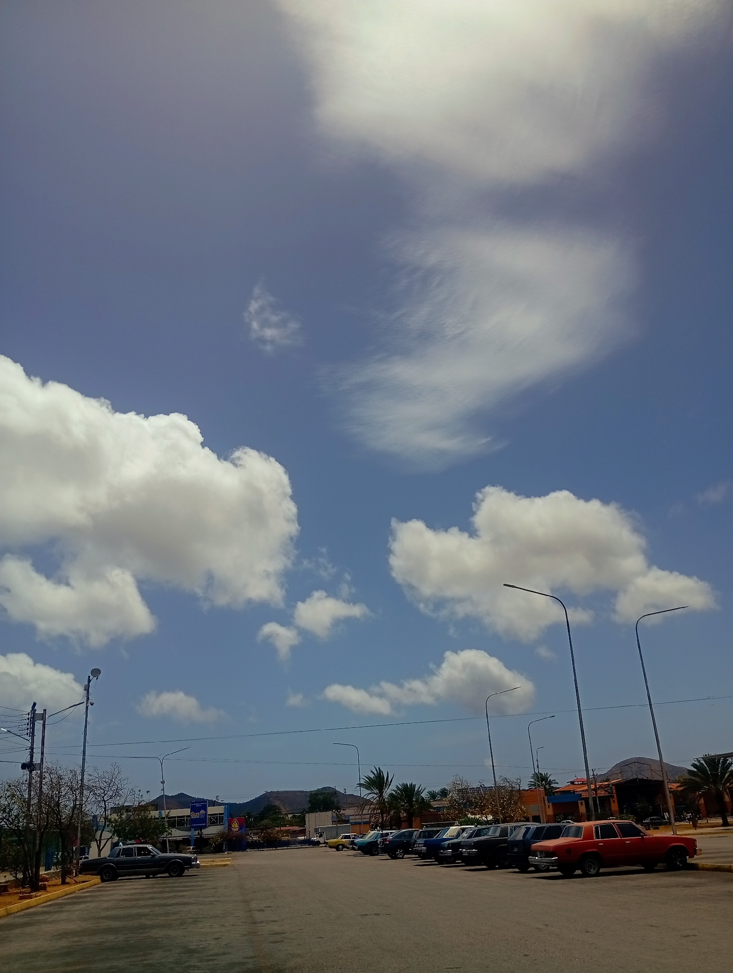 The image shows a wide asphalt parking lot with several cars parked in rows. The parking lot is situated in front of a low-rise commercial building with a blue facade and a prominent sign. In the background, mountains rise under a bright blue sky dotted with fluffy white clouds. Streetlights stand tall along the sides of the parking lot. The scene appears to be in a warm, sunny climate. The overall impression is that of a typical daytime scene in a commercial area. This location is identified as being in Juan Griego, Venezuela.