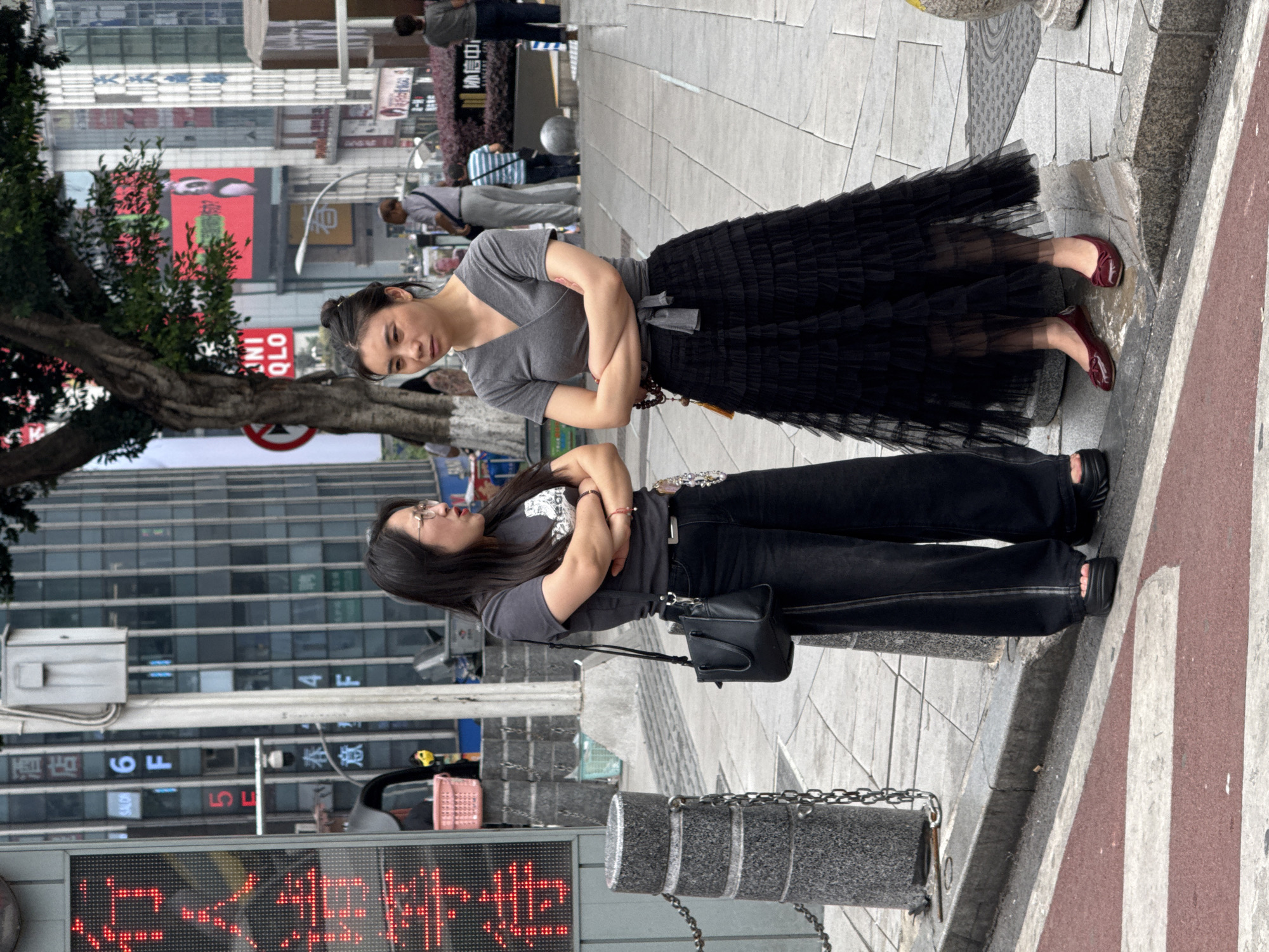 Two women are standing side by side on a sidewalk in an urban area of Liangjiang, China. The woman on the right is wearing a grey wrap-style top and a long, black, multi-layered tulle skirt. She has her arms crossed. Her shoes are dark red ballet flats. The woman on the left is also wearing a grey t-shirt and black jeans. She has a black handbag with a long strap. Her sandals are black. She is also standing with her arms crossed. A city street with buildings and digital signs and a tree can be seen in the background. The signage includes "UNI QLO". A traffic control device is partially visible above the woman on the right.