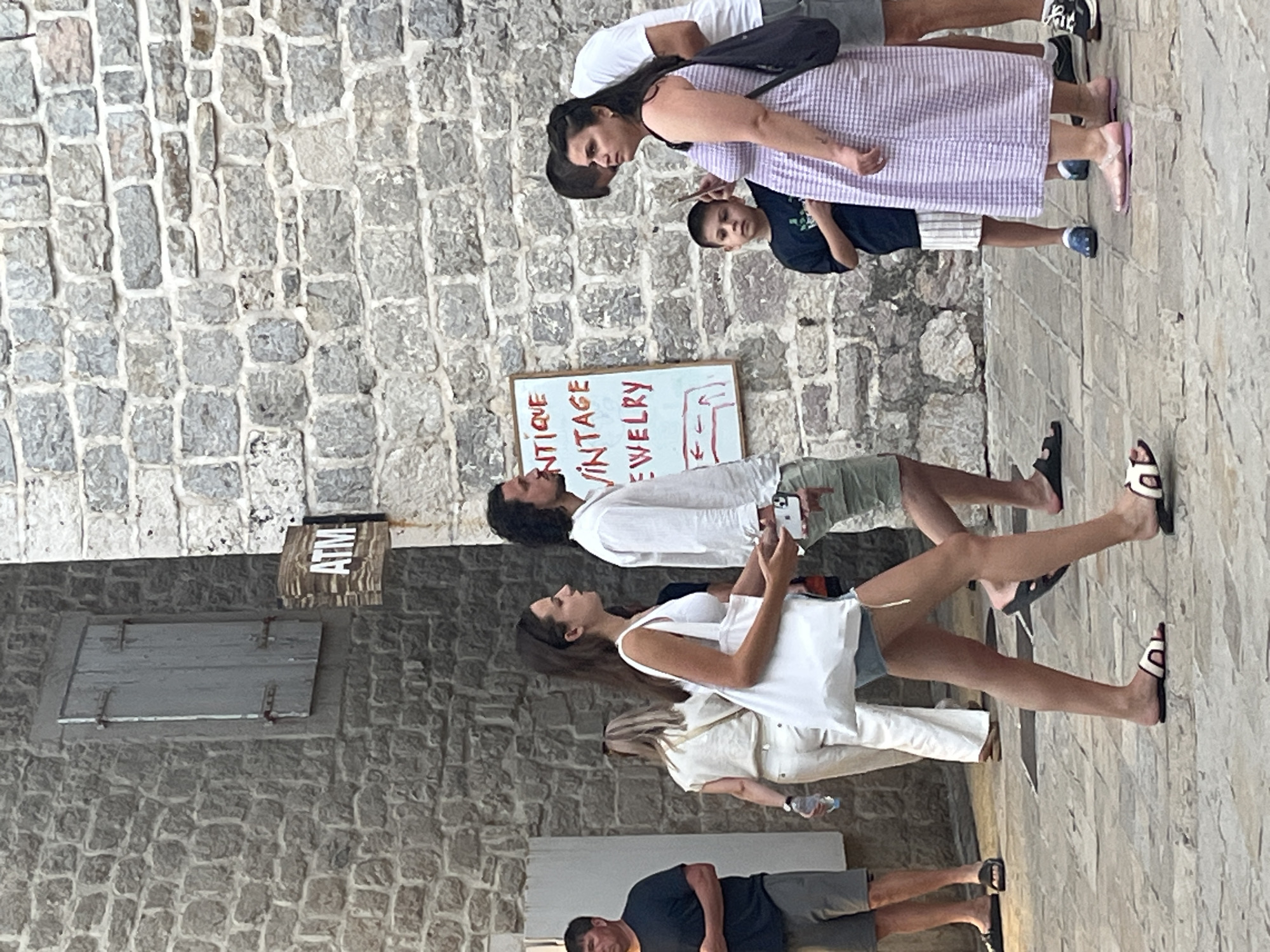 The image captures a street scene in Budva, Montenegro, characterized by its old town architecture. In the foreground, several people are walking on a cobblestone path. A woman in a white top and denim shorts, carrying a white tote bag, is walking towards the right. To her right, a woman in a light purple and white checkered dress is also walking. Behind them, a young boy in a dark t-shirt and striped shorts stands with a mobile phone. Further back, a man in a white linen shirt and light-colored shorts is walking, and to his left, a woman in a white top and pants. On the far left, a man in a dark t-shirt and gray shorts is standing.
The background features a textured stone wall, typical of the historic buildings in Budva. A wooden sign with the letters "ATM" is mounted on the wall. A white board sign with red lettering that reads "ANTIQUE VINTAGE JEWELRY" is also visible, along with directional arrows drawn in red. The overall lighting suggests it is daytime, with soft shadows indicating diffused sunlight. The scene depicts a moment of pedestrian activity in a historic urban setting.