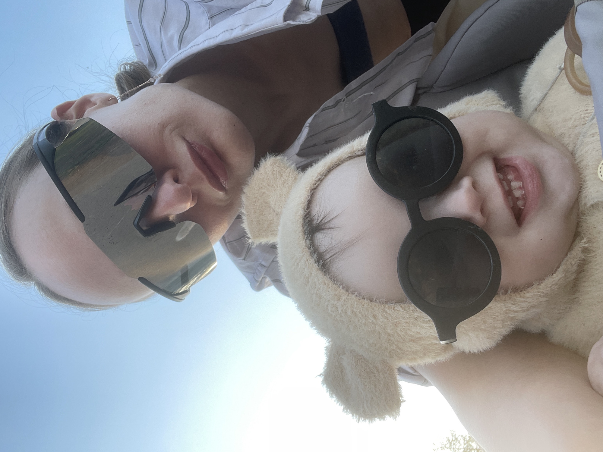 Two individuals are captured from a low angle perspective against a bright, clear sky. A woman with light skin and blonde hair, partially obscured by mirrored sunglasses, is wearing a striped shirt. Beside her, a child with fair skin is wearing a fuzzy, hooded outfit with bear ears and round, dark sunglasses, revealing a toothy grin. The image appears to be taken outdoors in natural daylight. The location, Подшивалово, Russia is not visually discernible from the image.