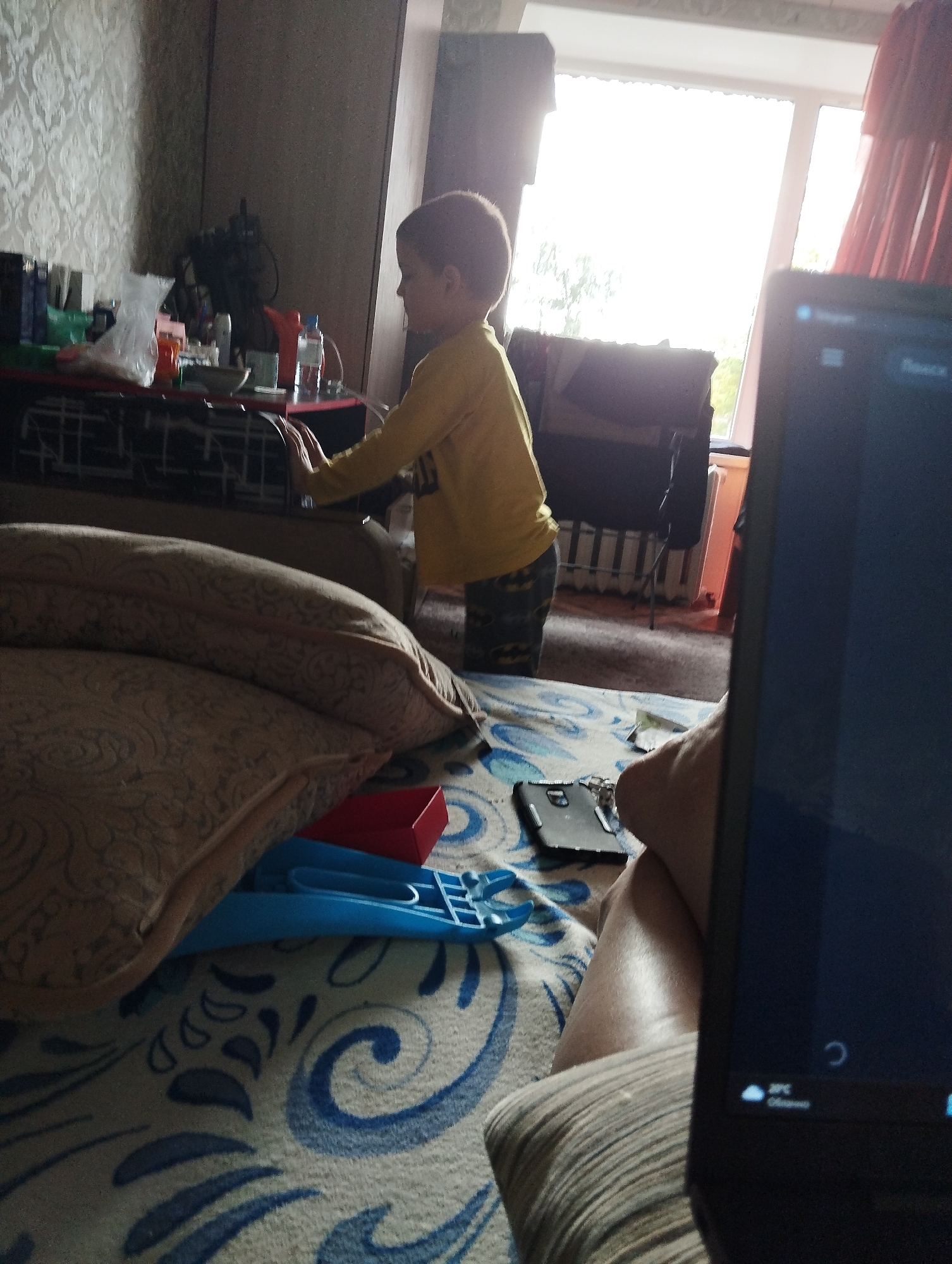 The image depicts an indoor scene, likely a living room or bedroom, with a young boy standing near a table. The boy is wearing a yellow long-sleeved shirt and patterned pants. He is facing away from the camera and appears to be reaching for something on the table. The table holds various items, including bottles, a plastic bag, and what looks like a bowl. In the foreground, part of a laptop screen is visible, showing a loading icon and temperature information. A couch with patterned cushions is also in the foreground. A window with red curtains is visible in the background, letting in bright daylight. The room has patterned wallpaper on one wall. The scene appears to be in Kingisepp, Russia, based on the provided context.