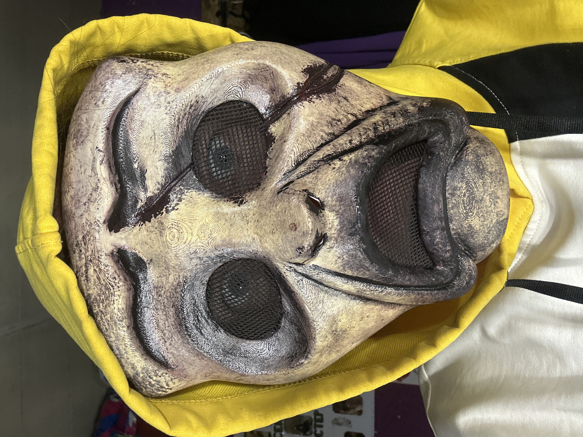 A close-up, high-angle shot captures a stylized, eerie mask partially nestled within a bright yellow hood. The mask, appearing to be made of a rigid, textured material, possibly 3D printed given the visible layer lines and wavy patterns across its surface, features a pale, off-white base color. Dark, smudged accents, resembling dirt, grime, or dried blood, are prominent around its large, meshed eye sockets and wide, unsettlingly grinning mouth. The mesh covering these openings is dark, likely black, adding to its mysterious and somewhat macabre appearance.
The yellow hood, made of fabric, frames the top and left side of the mask, contrasting sharply with its muted, stained tones. To the right and behind the mask, parts of a white, black, and yellow jacket or shirt are visible, featuring clear stitching and a dark strap. The background is blurred and indistinct, suggesting an indoor setting. Fine, hair-like strands are visible near the junction of the hood and mask in some cropped views. In the bottom right corner of the original image, faint, blurred Cyrillic text can be vaguely discerned on an object in the background, consistent with the location context of Saratov, Russia. The mask's expression conveys an unsettling, almost sinister cheerfulness.