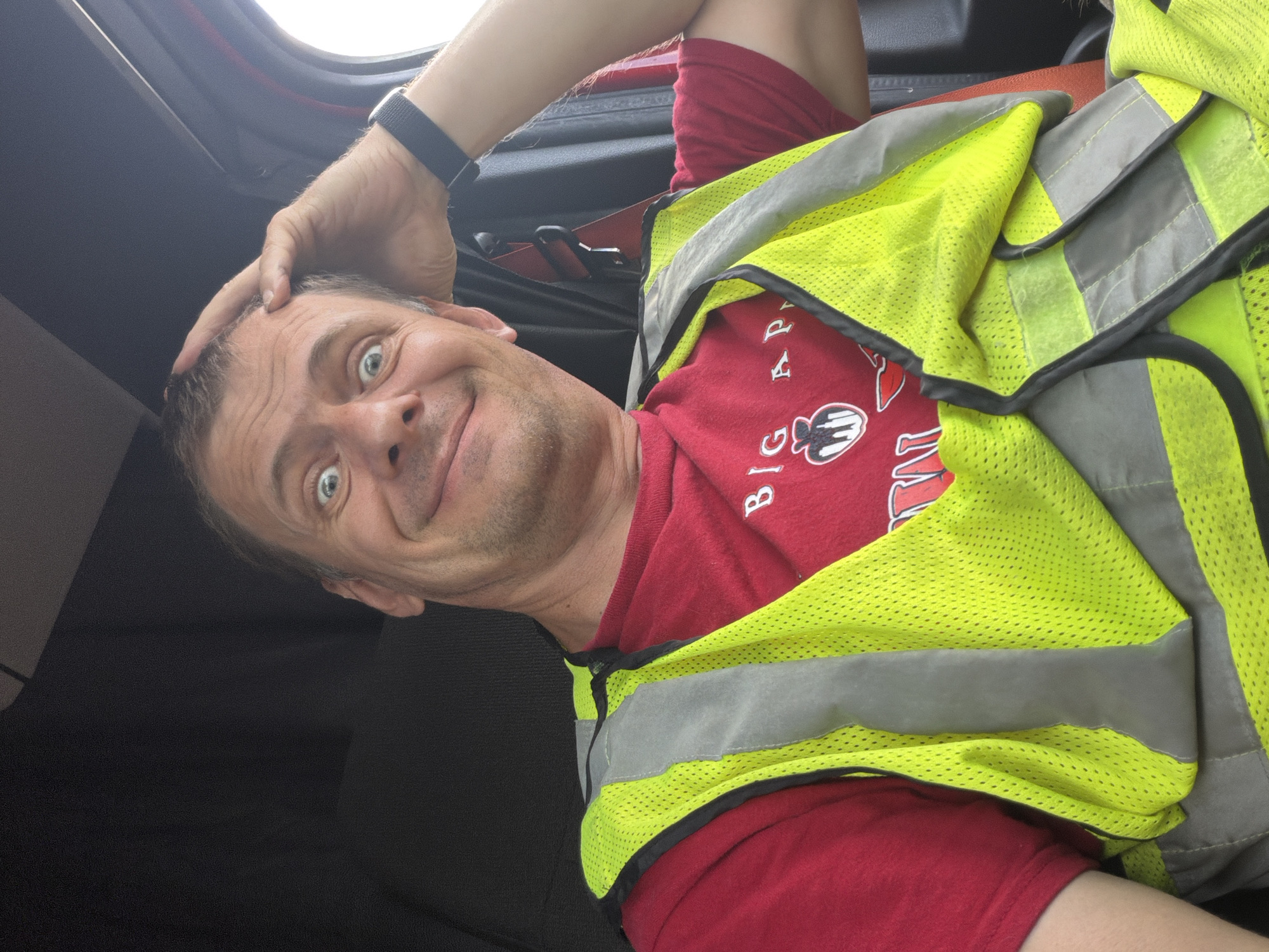 A man with a surprised expression and his hand on his head is captured in a selfie taken from inside a vehicle. He is wearing a red t-shirt with a graphic and text that reads "BIG AP..." and a bright yellow, reflective safety vest over it. His eyes are wide open, and his mouth is slightly open in a smile. The background shows the interior of a vehicle, with dark upholstery and a glimpse of a red seatbelt. The lighting suggests it is daytime. The image does not provide any specific visual cues to determine its location within the city of Chattanooga, United States of America.