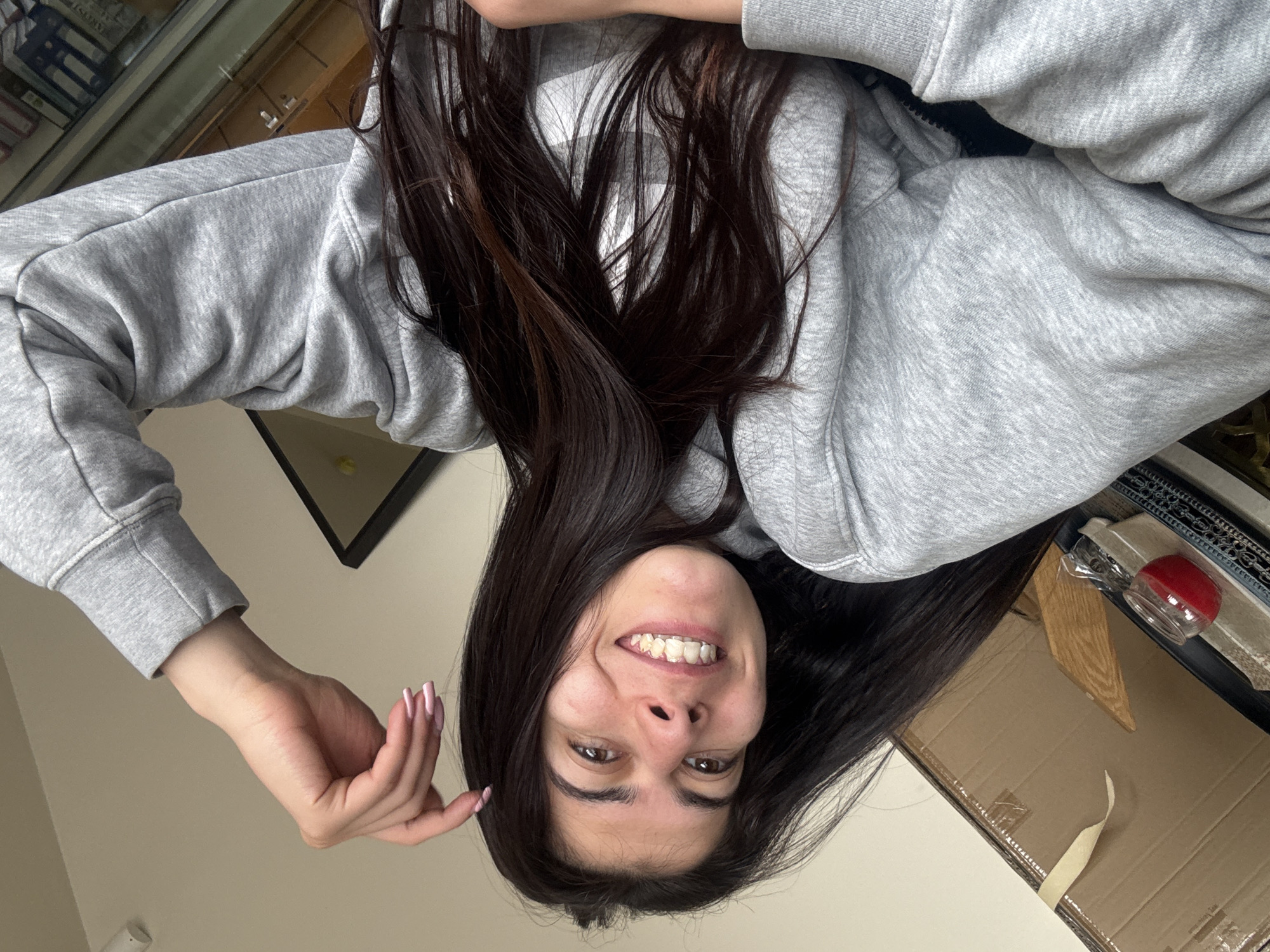 A young woman with dark hair and brown eyes, wearing a gray hoodie, is smiling at the camera. Her hand is raised near her face, and her nails are painted light pink. The background includes a window with reflections, a black framed picture, and a cardboard box with a red object on it. The location in Sofia, Bulgaria, cannot be determined from this image.