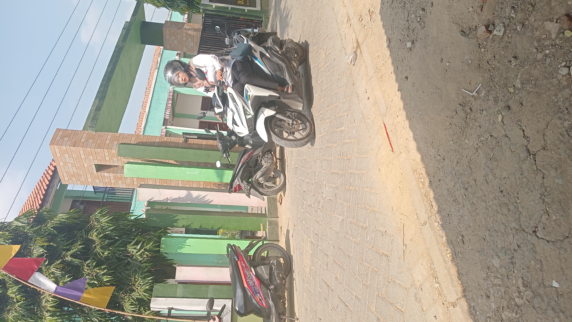 In Kedungwaru, Indonesia, a woman wearing a white long-sleeved shirt, black pants, and a light brown hijab is riding a white and blue scooter. She has a black helmet on. The scooter is parked on a paved area in front of a building with light green walls and a tiled roof. To the left of the scooter, a red and black scooter is parked. Behind these scooters, a black and silver scooter is also visible. The building's facade features colorful vertical pillars in shades of light green, pink, and white. A flag with yellow, red, and purple segments is attached to a pole on the left side of the image. The ground in front of the building is paved with rectangular bricks, and further in the foreground, there is a rougher, unpaved area with dirt and small stones. A shadow is cast across the unpaved ground from the right.