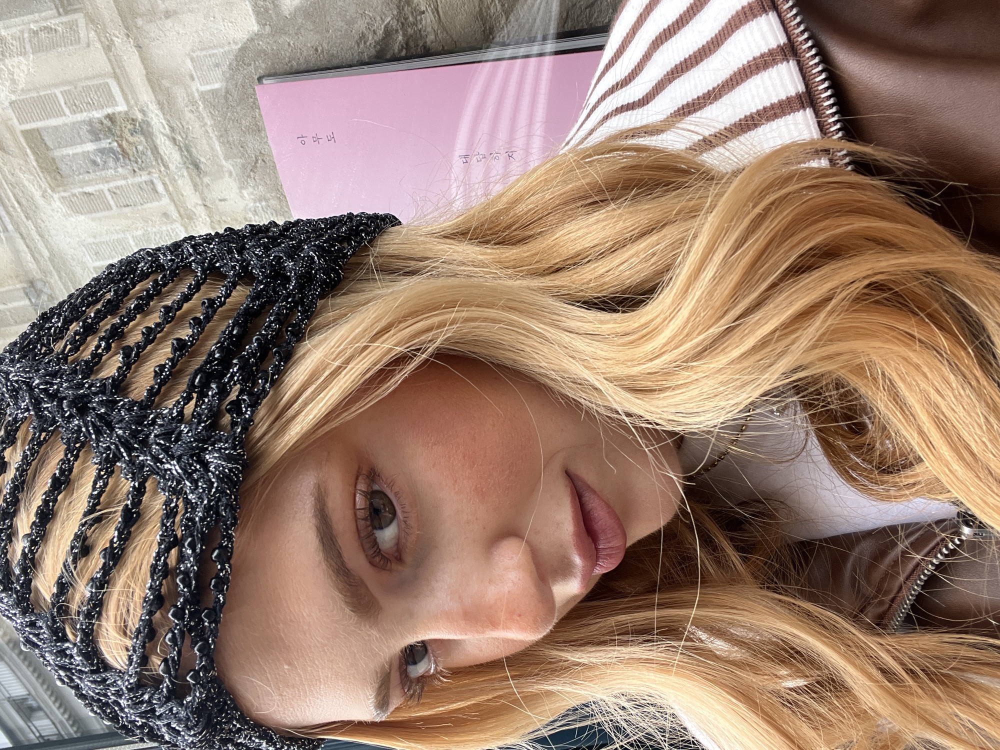 A close-up, slightly angled, and rotated image shows a fair-skinned woman with long, blonde wavy hair wearing a black, crocheted head covering embellished with beads. The woman's face is partially visible, revealing her left eye and part of her nose and lips. She has light brown eyebrows and lashes, and her lips appear to have a slight sheen.
Behind her head, there's a pale pink rectangular object, possibly a book or a folder, with some text in what appears to be Korean or a similar East Asian language. Part of a brown leather jacket with a zipper and a white and brown striped garment is visible.
The background is a muted gray, possibly a concrete or stone surface. It is difficult to infer the location as Paris, France, as there are no clear identifiable landmarks or environmental cues that would indicate the city.