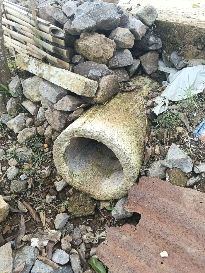 The scene in Đăk Mar, Vietnam, depicts an outdoor area filled with various debris and natural elements. A weathered, cylindrical concrete pipe section, showing significant moss or algae growth, lies horizontally in the midground, its interior visible. Directly behind and to the left of this pipe, a substantial pile of diverse rocks and stones is accumulated. Integrated into the lower section of this rock pile is a segment of a bamboo fence or screen, horizontally supporting a rectangular concrete beam. In the lower right foreground, a piece of severely rusted, corrugated metal sheet is visible. The ground is composed of loose soil, small stones, dried leaves, and sparse green vegetation, including grass shoots. A crumpled white plastic bag is partially visible near the right edge. A tree trunk is partially visible on the far left. No individuals are present, and no actions or interactions are occurring.