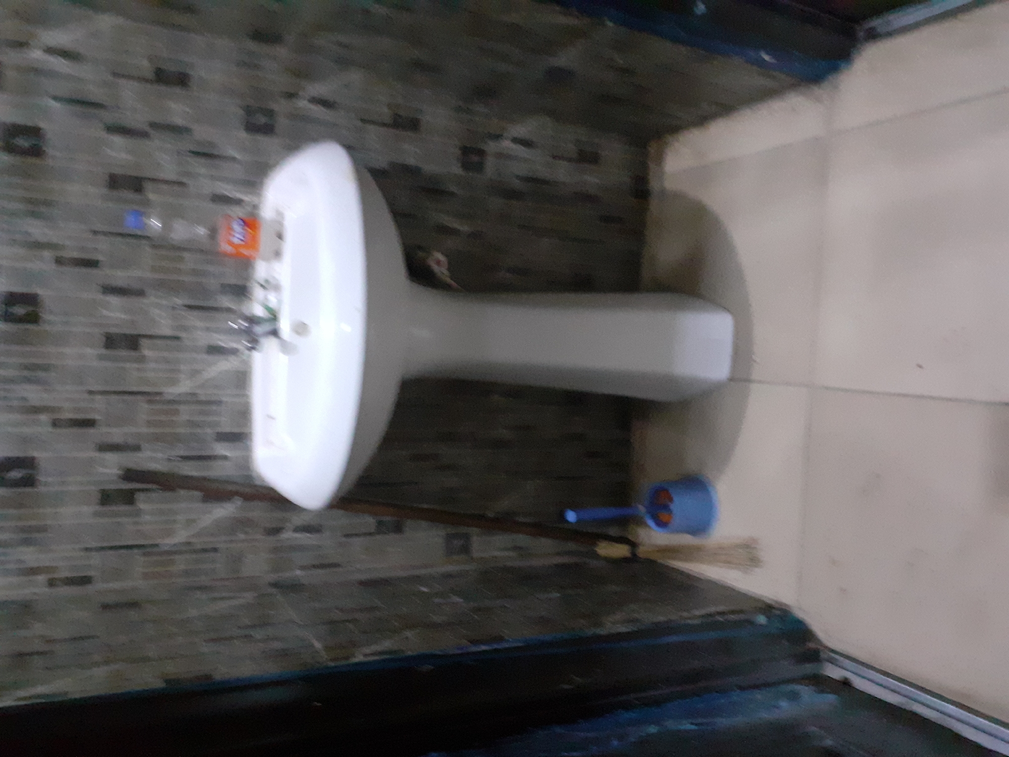 The image shows a bathroom in Jalingo, Nigeria. A white pedestal sink with a chrome faucet is the central focus, with a partially visible bottle of Fanta and another drink nearby. A blue toilet brush and holder are positioned on the floor to the left of the sink, next to a broom. The walls are tiled with a mosaic pattern of rectangular tiles in various shades of grey and brown. The floor is tiled with large, light-colored square tiles. The lighting appears to be artificial, casting shadows and giving the scene a somewhat dim ambiance. There are no people or animals visible in the image. The overall impression is of a functional but somewhat cluttered bathroom.