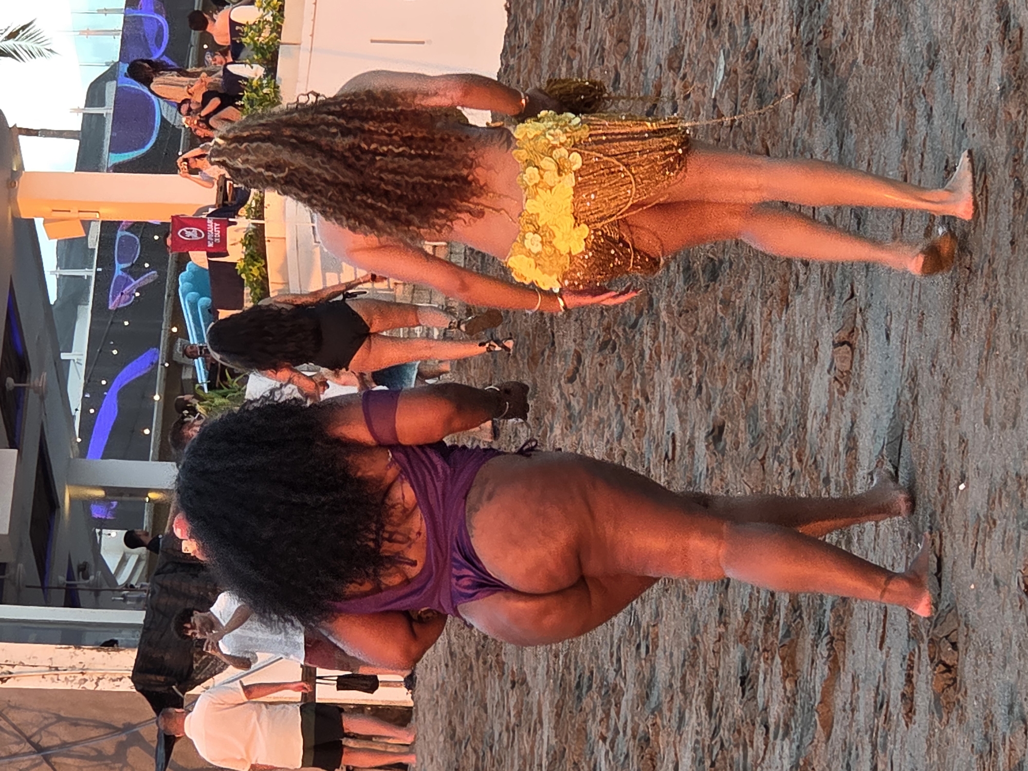 Two women walk on a sandy beach in North Kuta, Indonesia. The woman on the left, who has dark curly hair, wears a purple bathing suit. She is walking away from the camera. The woman on the right, who has long, curly blonde hair, wears a yellow, sequined skirt and a light-colored top. She is also walking away from the camera. Other people are visible in the background, along with some buildings. The lighting suggests it is late afternoon or early evening.