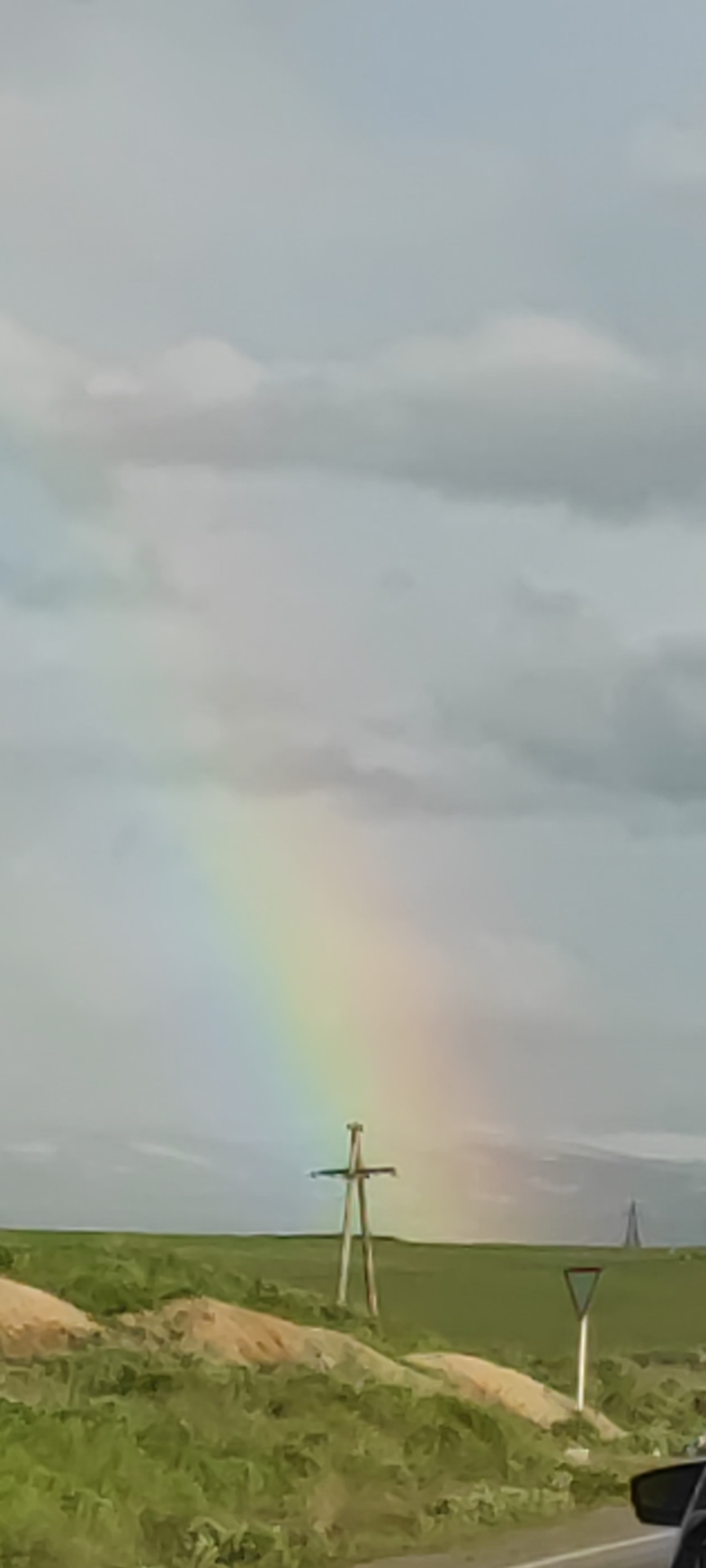 The image shows a landscape near Azatan, Armenia, featuring a partial rainbow in the sky. The landscape includes green fields, small embankments of dirt, a yield sign, and a wooden utility pole in the middle ground. A distant, tall structure is visible on the horizon. Part of a vehicle's interior (specifically, a side mirror) is visible in the lower right corner of the image. The sky is overcast with gray and white clouds.