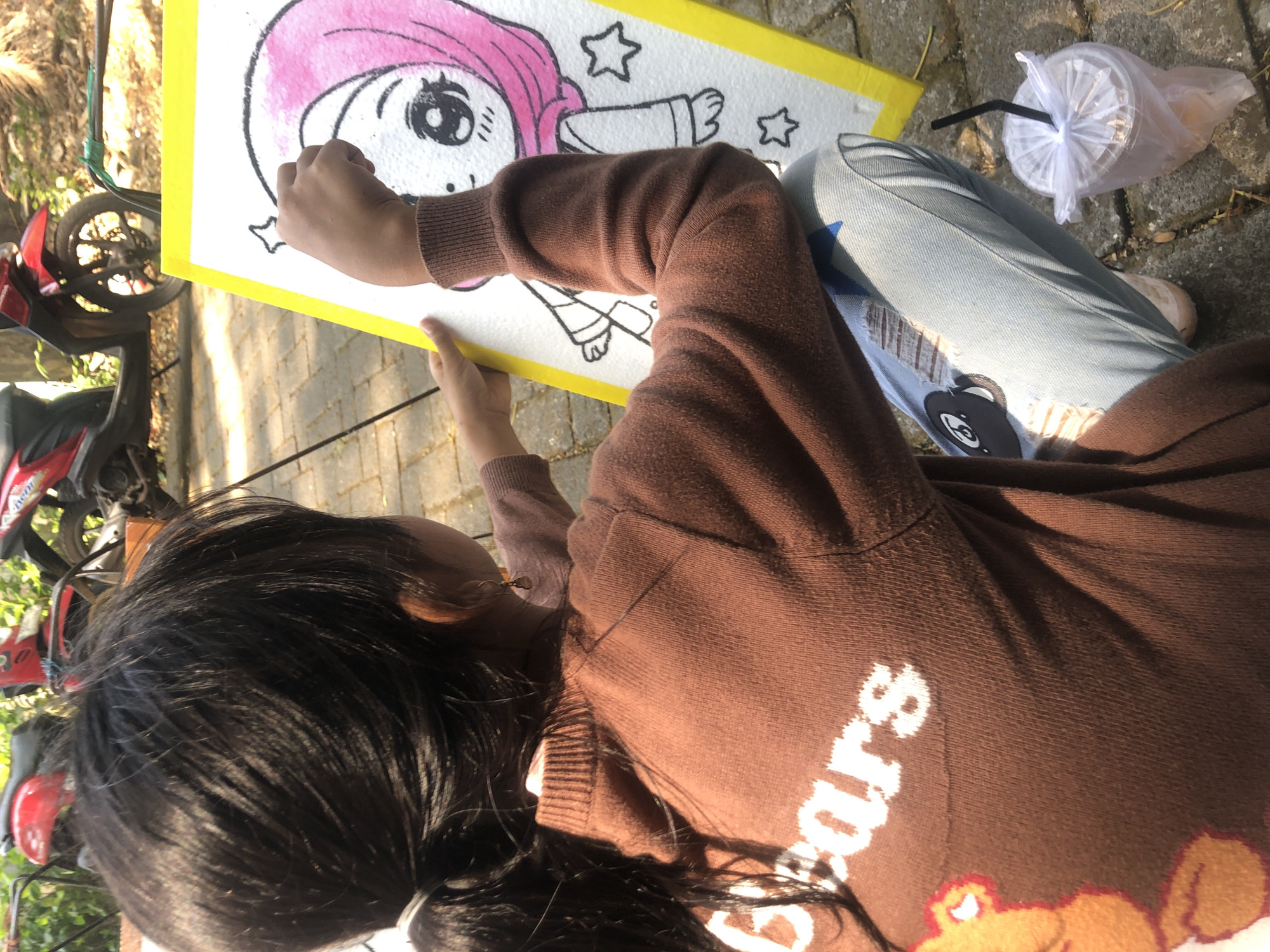 A young girl, with dark hair pulled into a ponytail and wearing a brown sweater with "Bears" printed on it in white, sits outdoors and draws on a white canvas on an easel. The canvas has a cartoon drawing of a girl in a pink hijab. The girl's right hand is holding a drawing utensil, and she appears to be adding details to the drawing. She is wearing light blue jeans with patches. To the right of the girl, there is a plastic cup with a drink and a black straw, covered with a clear plastic bag. The background shows red motorcycles and foliage, suggesting an outdoor setting, possibly a cafe or a park in Pasuruan, Indonesia, given the context. The ground is paved with stone.