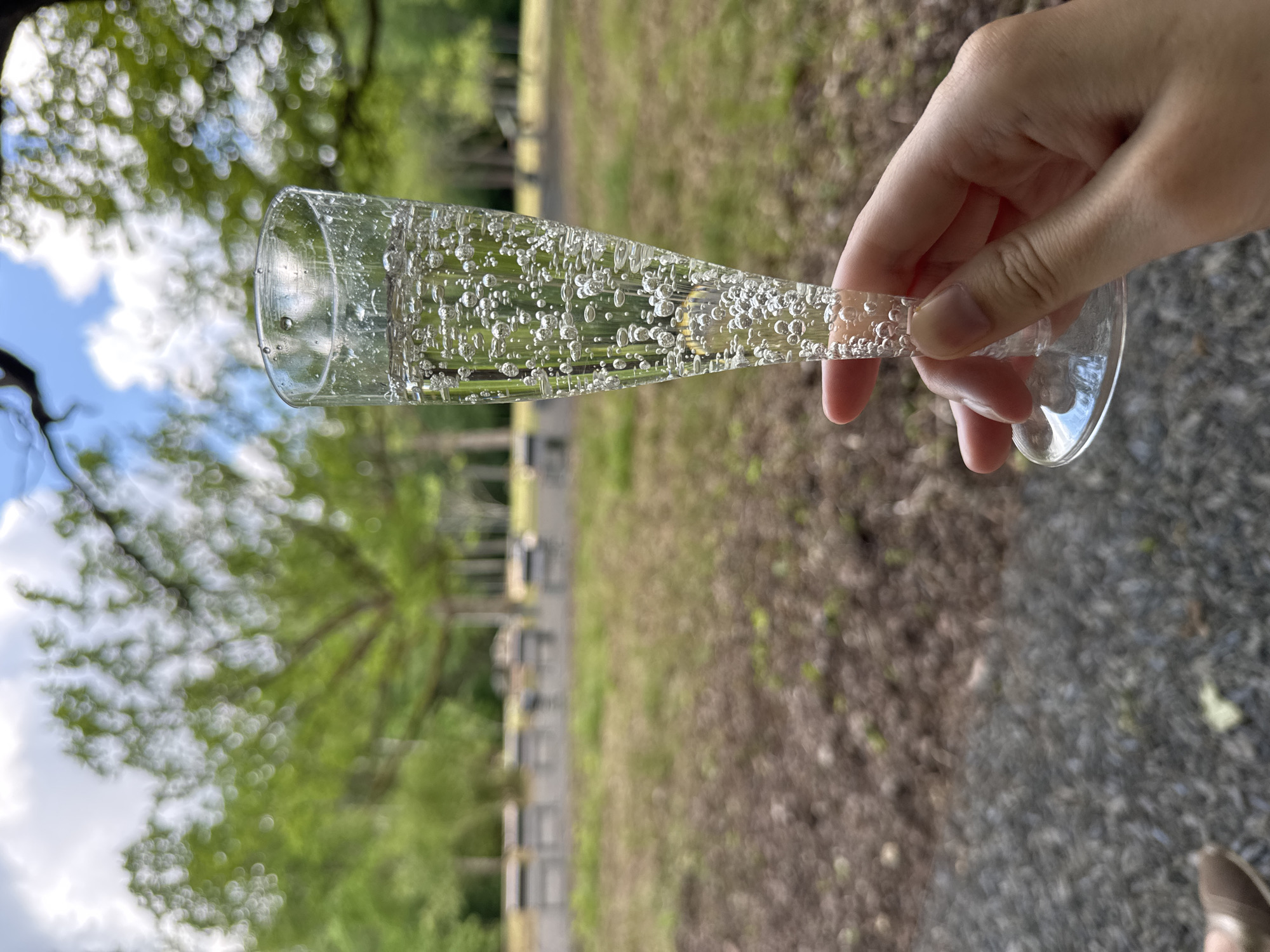 An extreme close-up shot captures a hand holding a flute glass filled with a bubbly, clear liquid, likely champagne or sparkling wine. The glass is tall and slender, with numerous small bubbles rising within the beverage. The background is blurred, suggesting an outdoor setting with trees and a bright sky. The ground in the foreground is covered with gravel. The overall impression is one of a leisurely moment, possibly a picnic or outdoor celebration. The image is not specifically set in Jūrmala, Latvia, and lacks any details to identify it as such.
