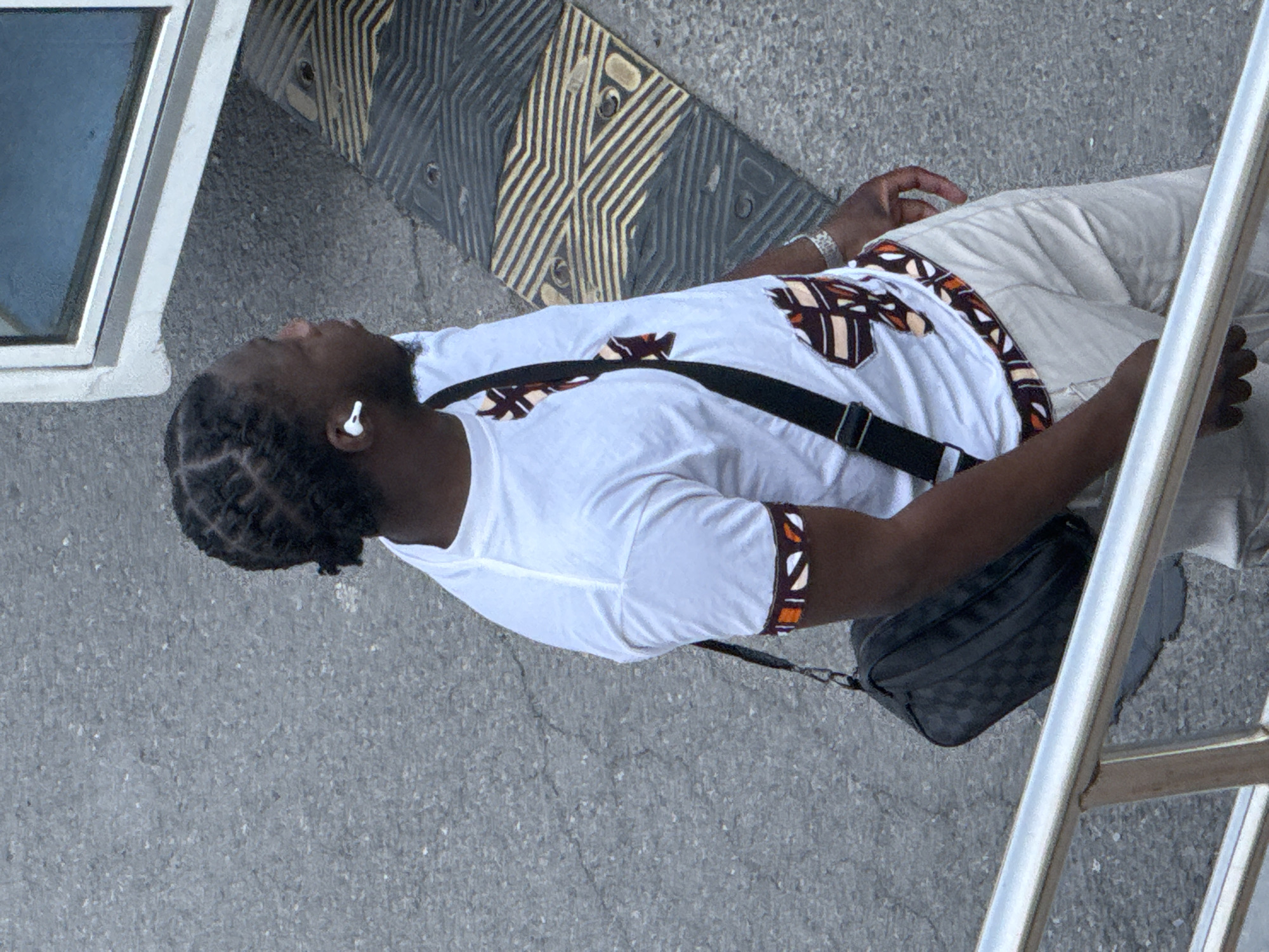 A man with dark skin and braided hair is outdoors on a gray concrete surface. He wears a white t-shirt with patterned brown trim, light-colored pants, and a black shoulder bag. An earbud is visible in his ear. There are architectural elements including a window frame and a metal railing. A speed bump or similar object with alternating stripes is visible in the background. The location is not determinable as Zhongtun, China.
