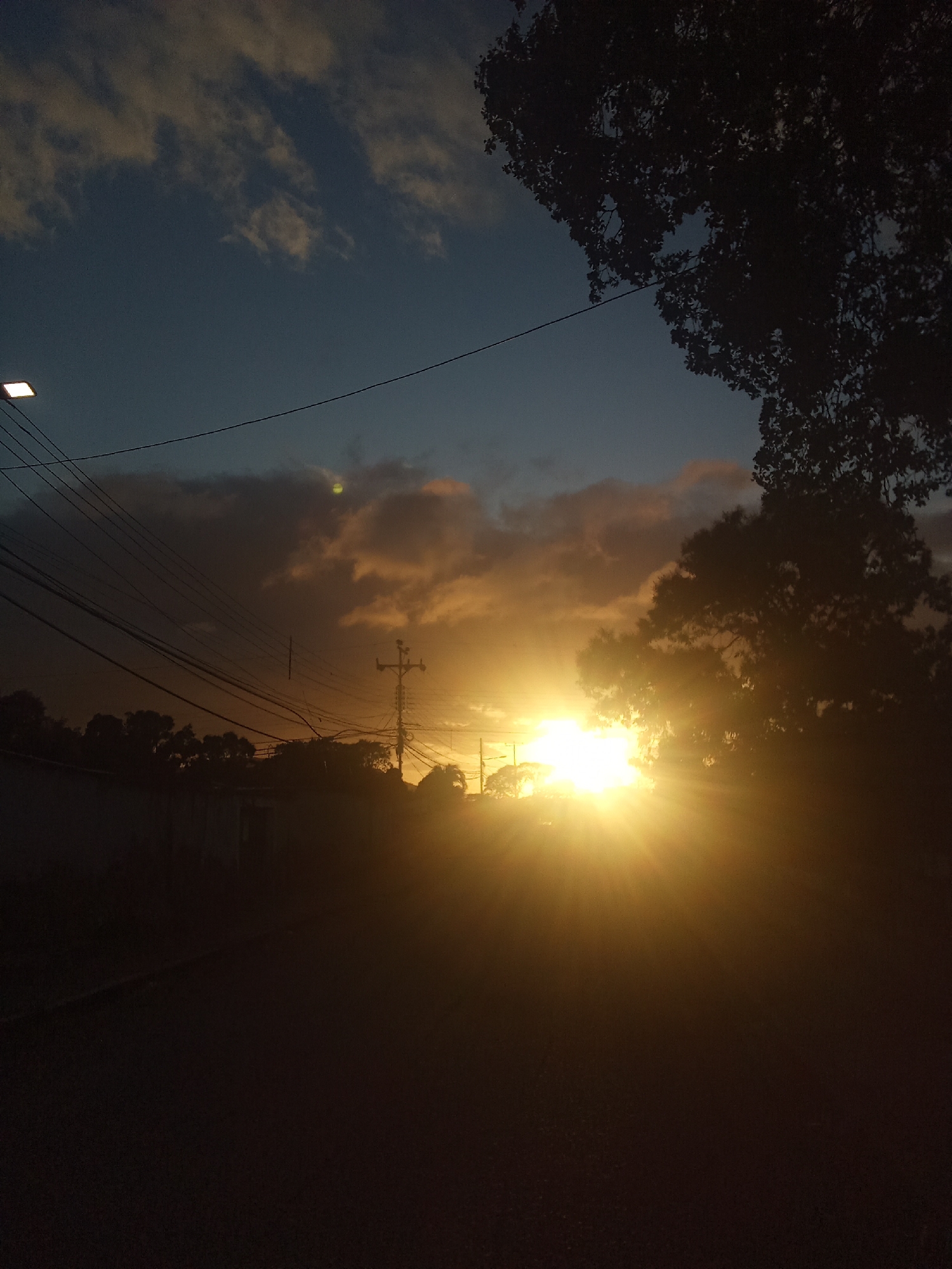 Captured at Juan Griego, Venezuela, the image depicts a sunburst dominating the horizon. In the foreground is a dark, indistinct road. Above, power lines stretch across the sky towards the sun. Silhouetted trees frame the right side of the image, while more trees and buildings create a dark band along the horizon. Clouds tinged with orange surround the sun, contrasting with the darker blue portions of the sky. A lamppost is visible on the left.