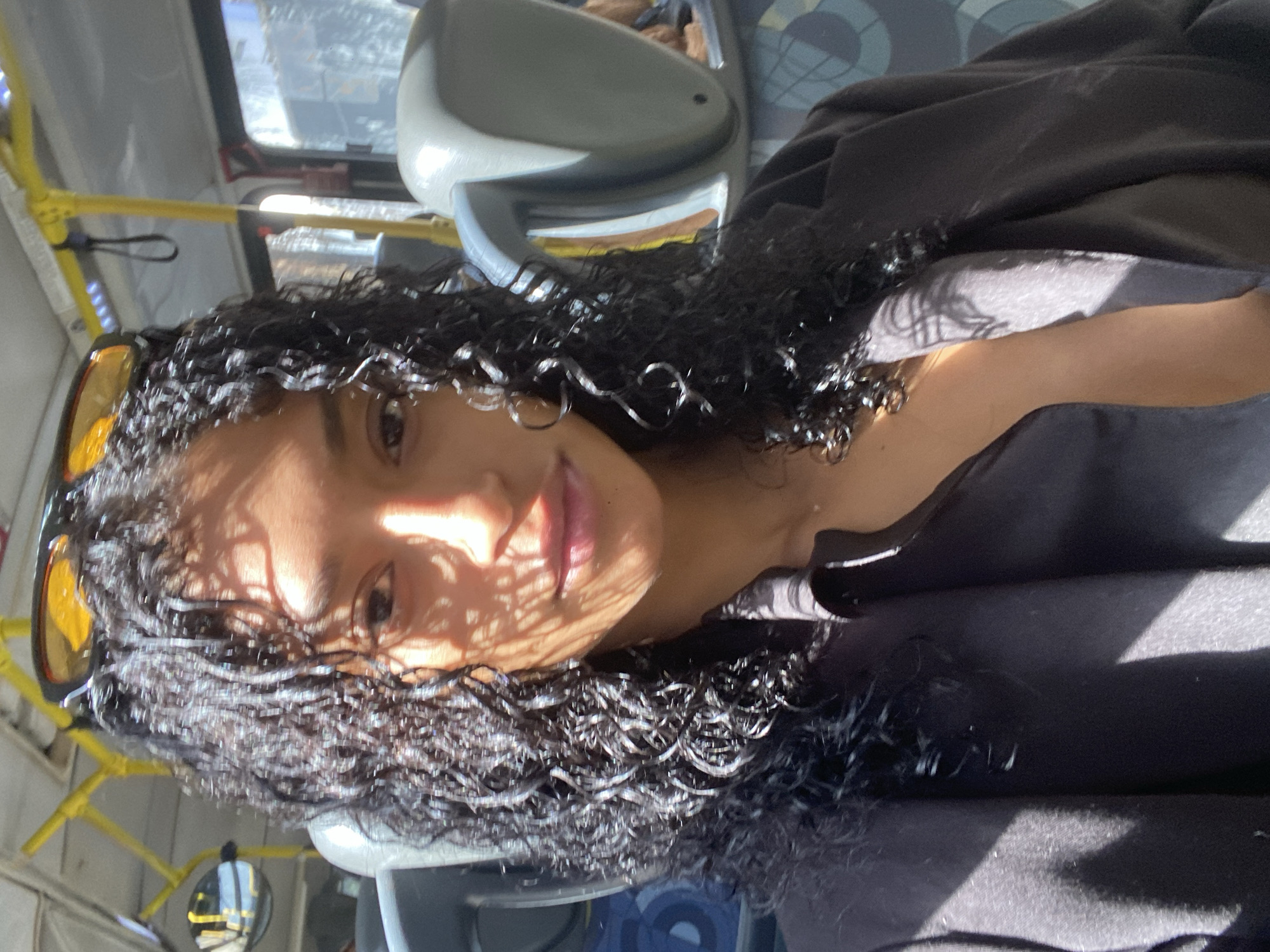 A selfie taken on a bus in São Paulo, Brazil. The person has curly black hair, dark skin, and is wearing a dark shirt. Sunglasses are perched on their head. Light streams through the bus windows, creating shadows on their face. The bus interior is visible, including seats and handrails.