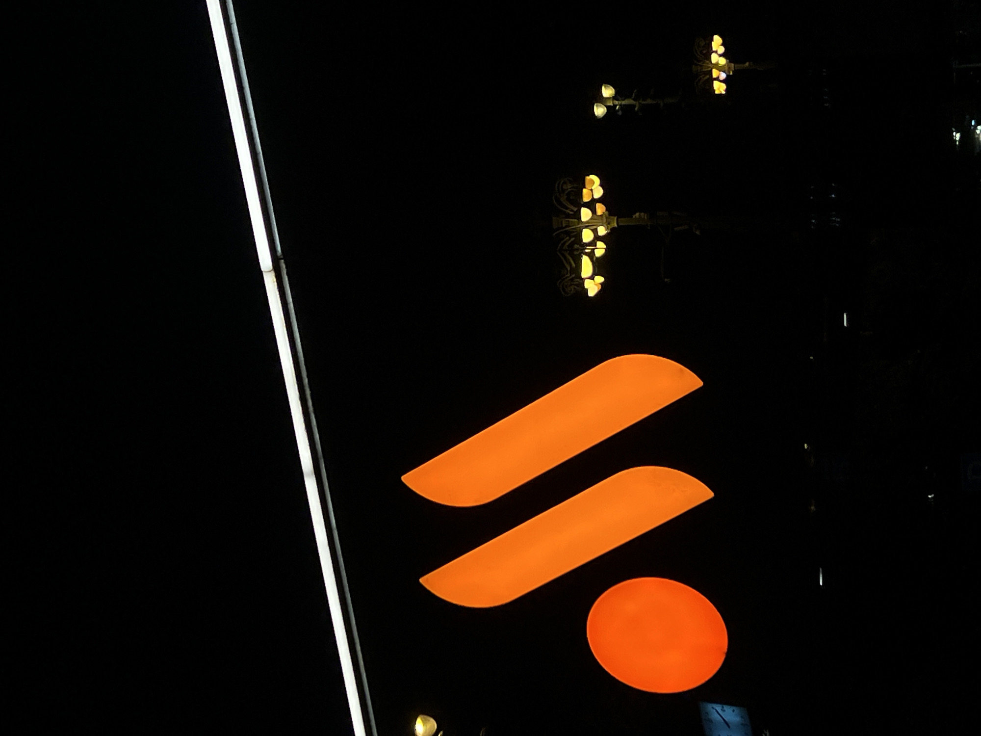 The image shows a night scene in Moscow, Russia. The illuminated orange logo of Sistema PJSFC, a Russian conglomerate, is visible on a dark background. A vertical white light fixture is situated to the left of the logo. In the background, streetlights are blurred and reflected, indicating a wet surface. No people are visible.