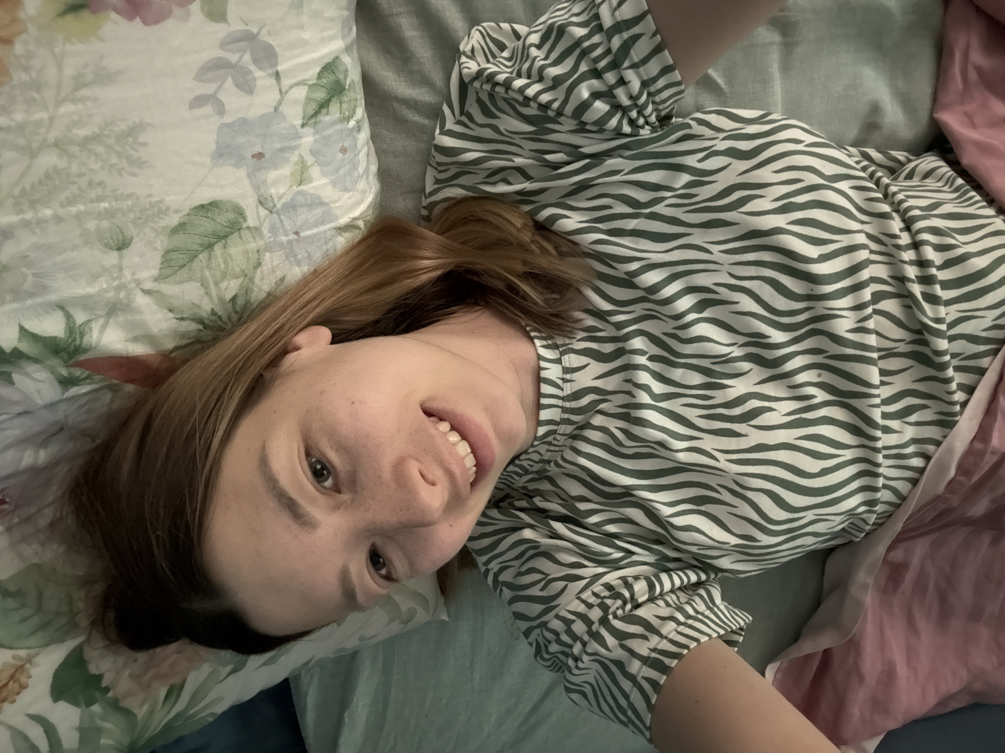A woman with light brown hair is lying on a bed with a floral patterned pillow and a green zebra-striped top. She is smiling and looking at the camera. The room appears to be a bedroom in Moscow, Russia.