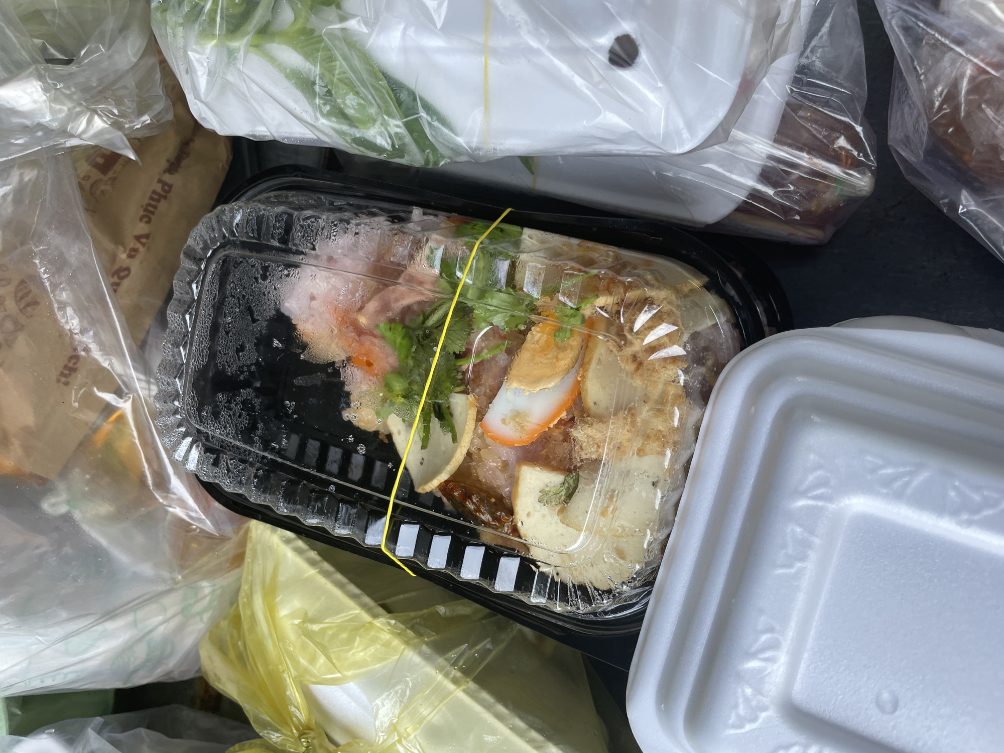 A close-up, overhead shot shows a takeout container of food secured with a yellow rubber band, nestled among other food containers and plastic bags. The takeout container has a clear plastic lid and a black base, and it contains what appears to be noodles with meat, egg, and herbs. The container is surrounded by white styrofoam containers, some wrapped in plastic. One plastic bag in the background has Vietnamese writing on it, which translates to "Phuc Thuy Ward". The scene appears to be in an outdoor setting, possibly a street food stall or a market. There are no people visible in the image.